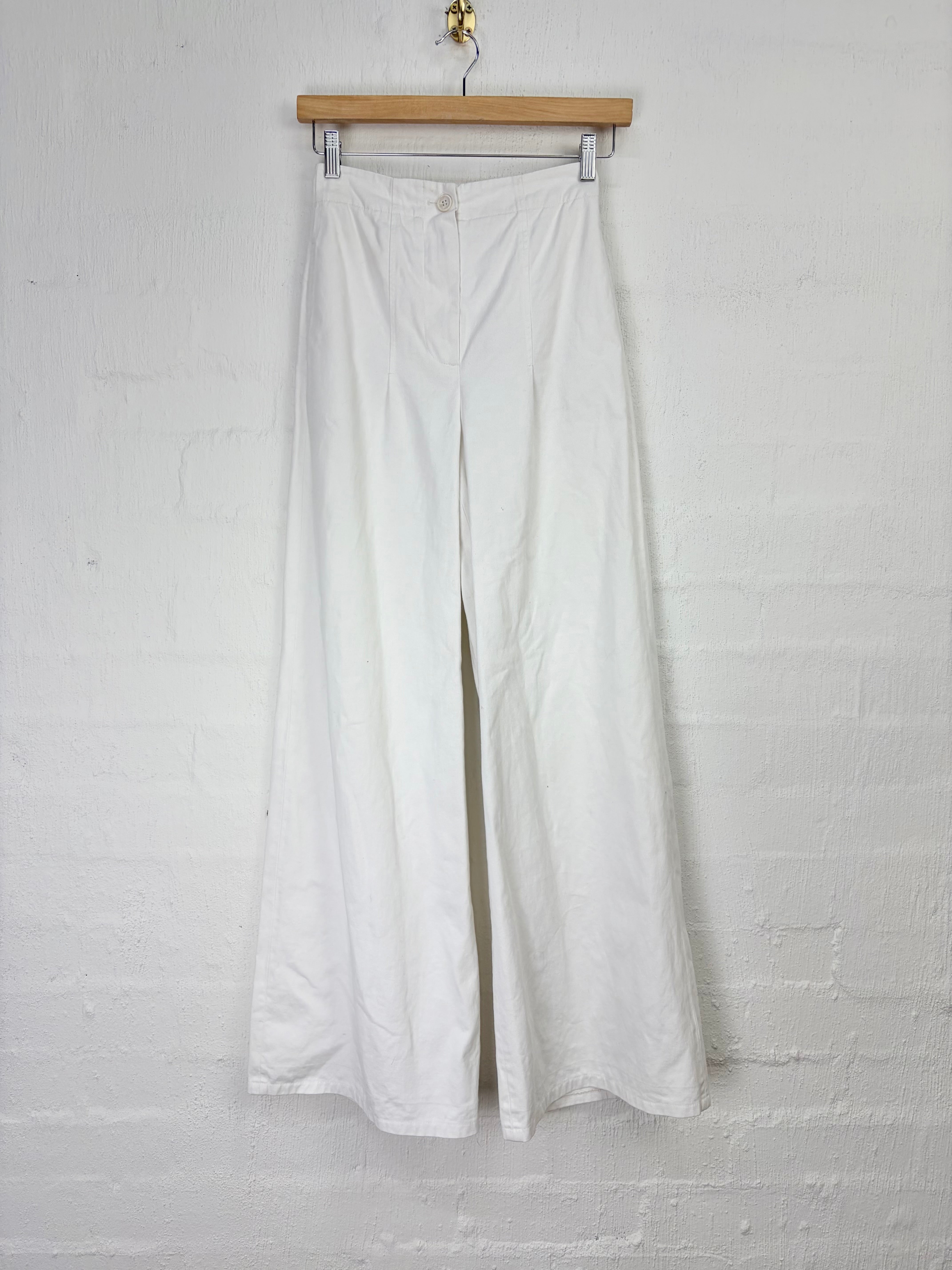 White wide-leg trousers hanging from a wooden hanger against a white painted brick wall.