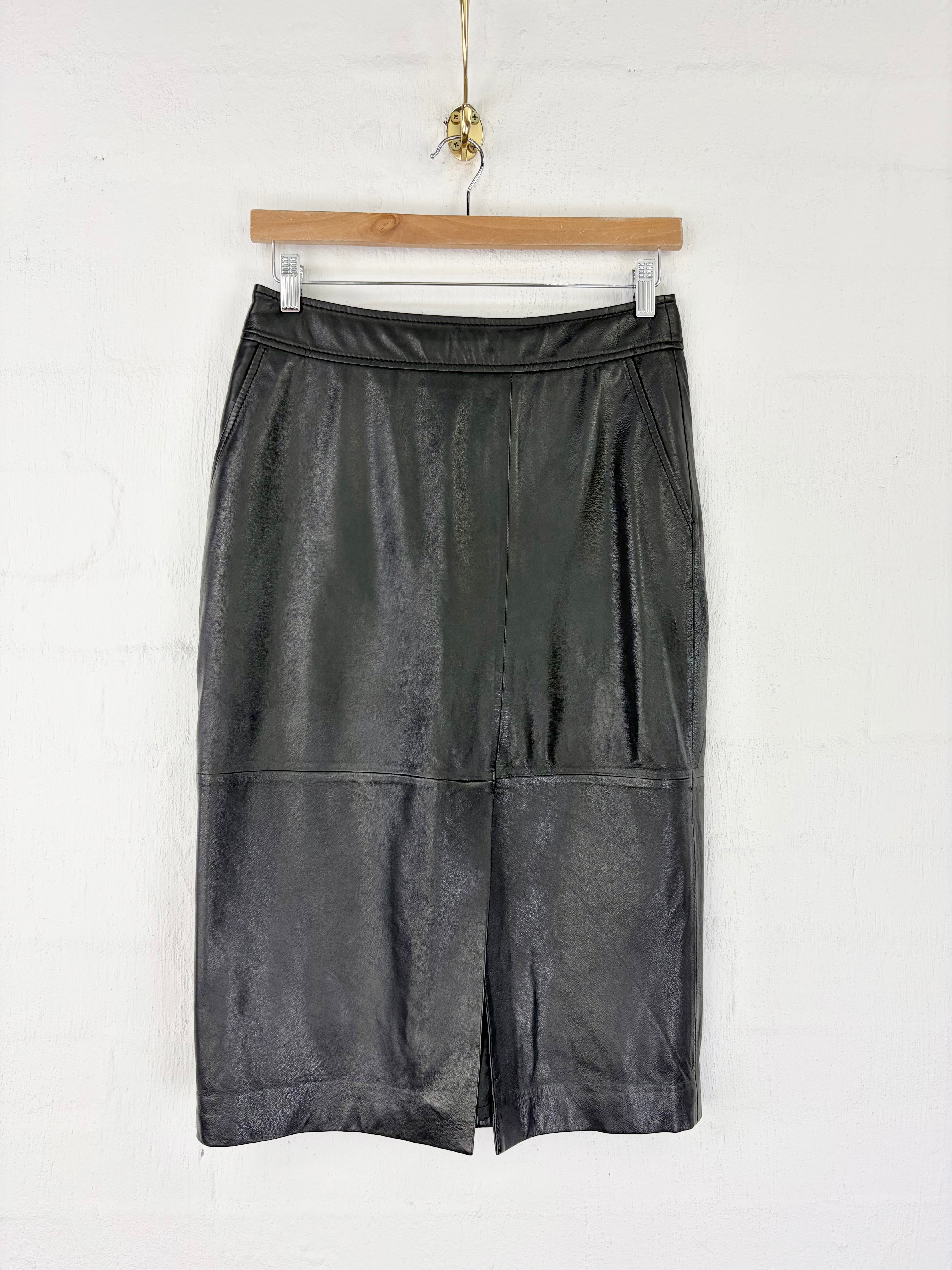 A black leather midi skirt with a front slit, hanging from a wooden hanger against a white wall.
