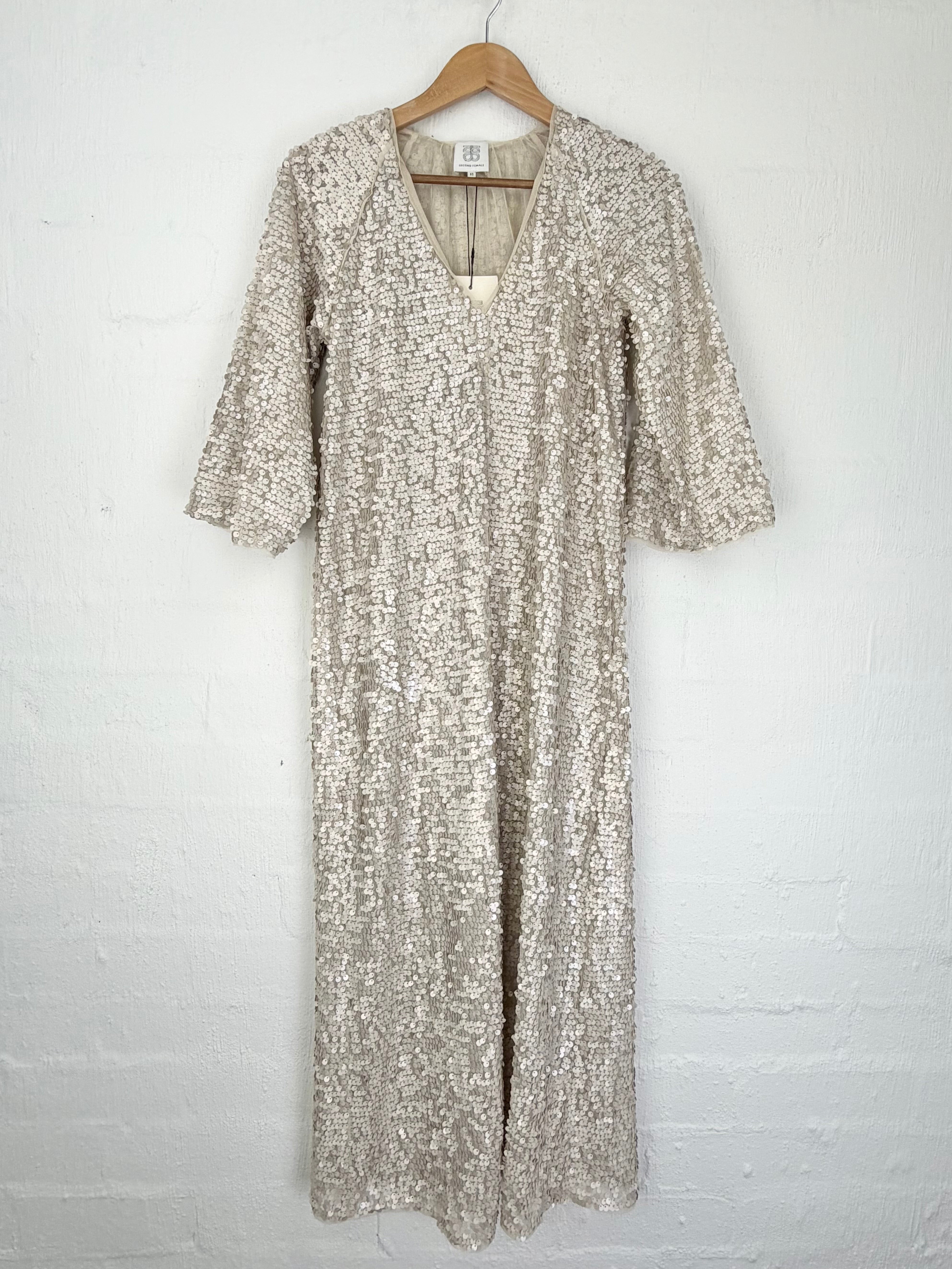 A long-sleeved, V-neck maxi dress covered in shimmering silver sequins, hanging on a wooden hanger against a white painted brick wall.