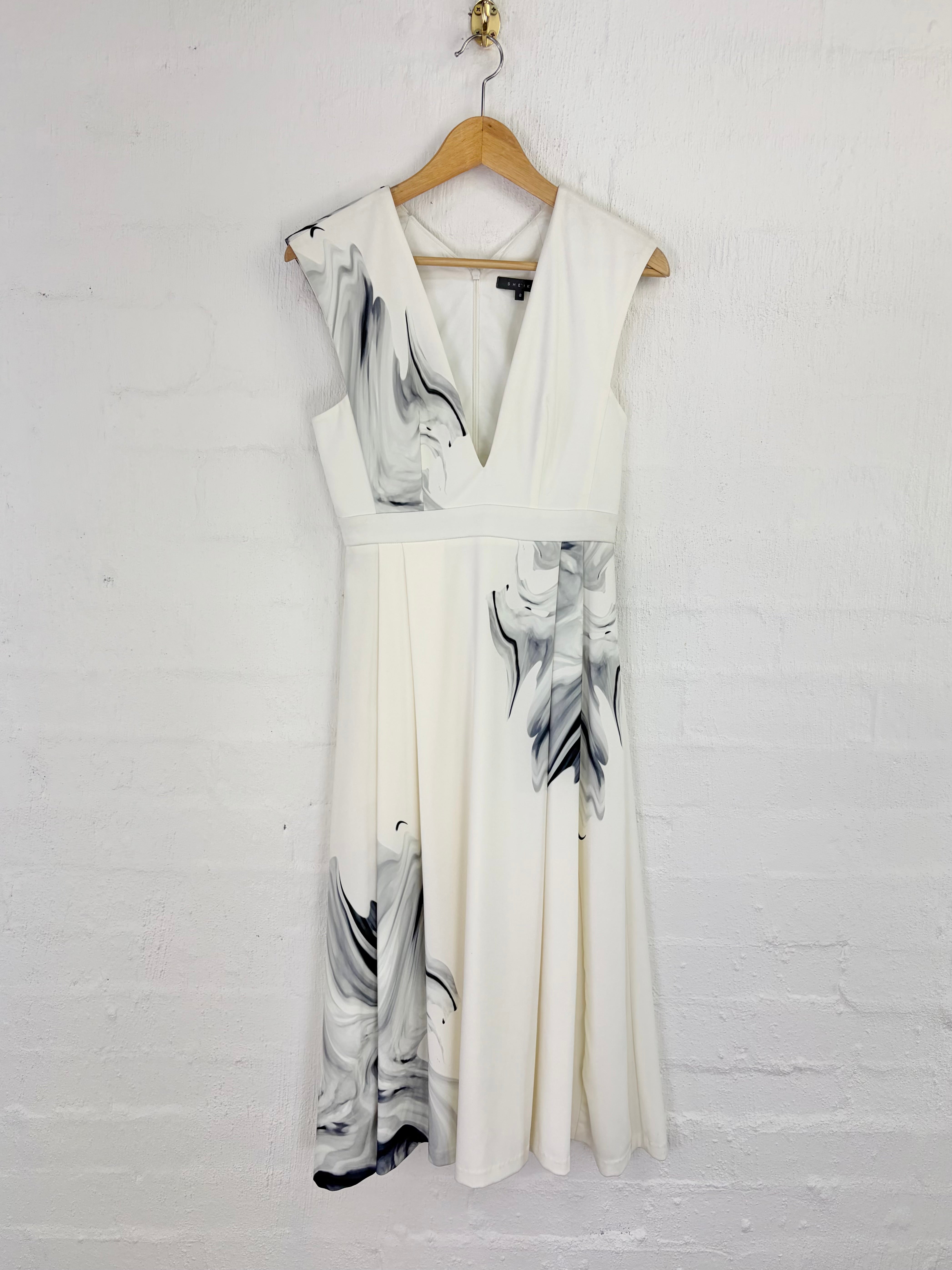 An off-white sleeveless V-neck dress with a black and gray marble print, hanging on a wooden hanger against a white painted brick wall.