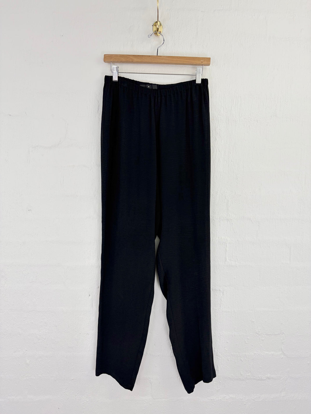 Black wide-leg cropped pants hanging from a wooden hanger against a white painted brick wall.