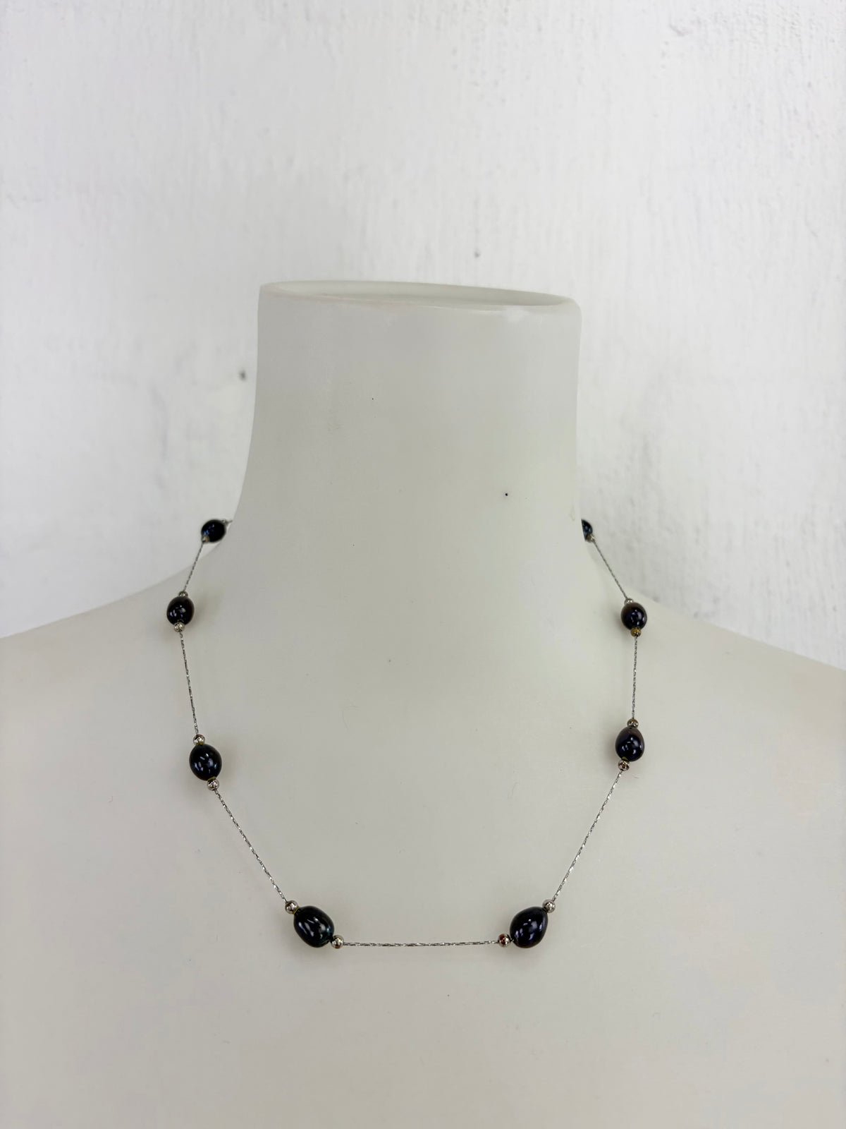 A delicate silver-toned necklace with small dark beads and a central horizontal bar with three larger dark teardrop beads, displayed on a white mannequin bust against a white wall.