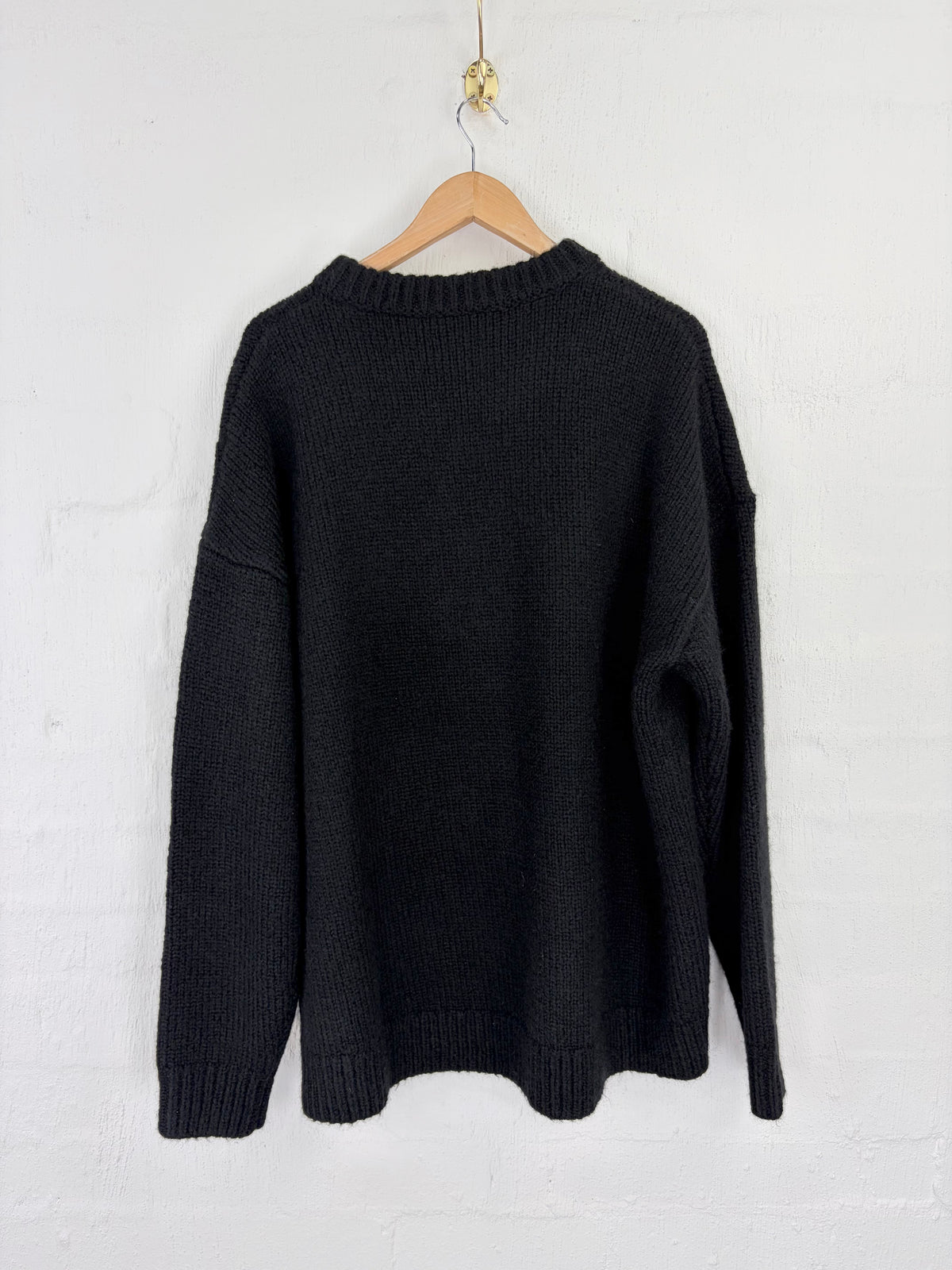 A black ribbed knit sweater hanging on a wooden hanger against a white painted brick wall.