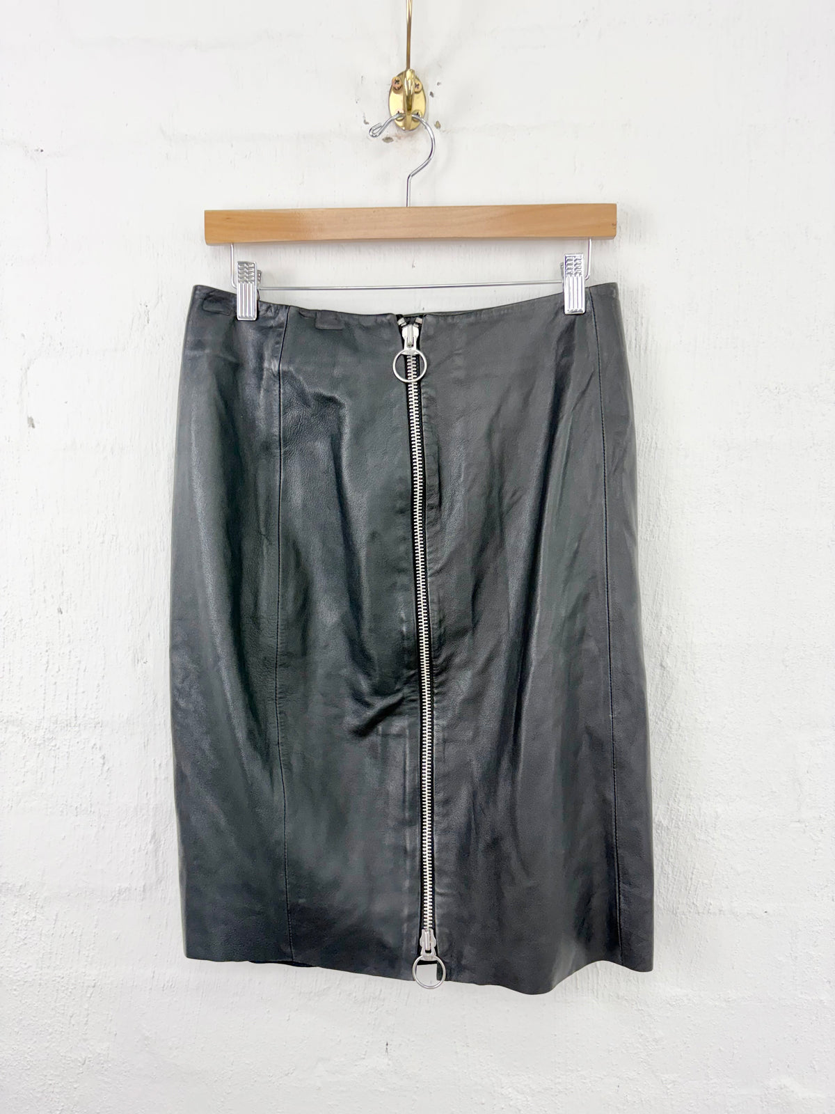 A black faux-leather pencil skirt with a prominent silver zipper down the front, hanging from a wooden hanger against a white painted brick wall.