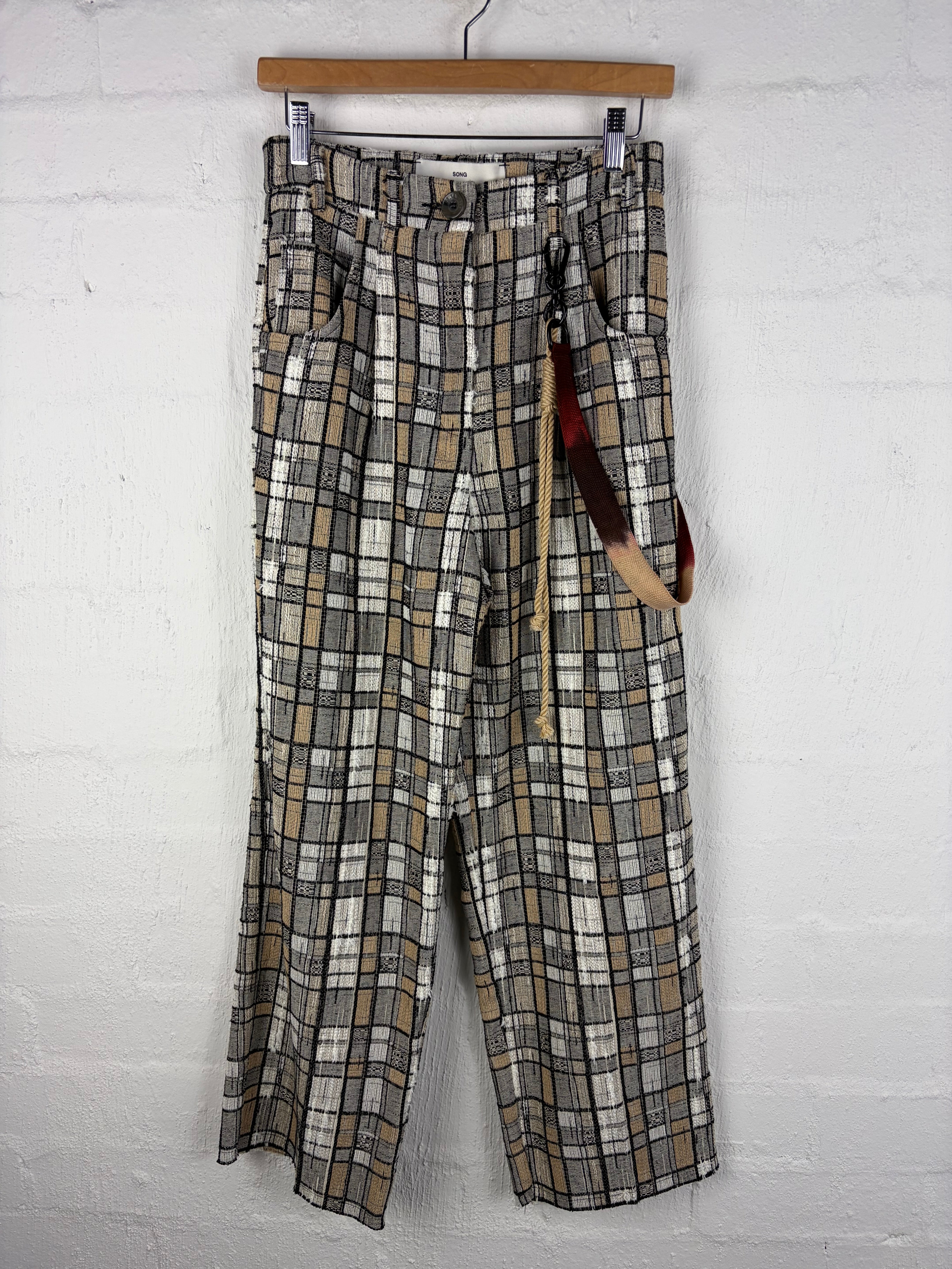 A pair of plaid trousers in beige, gray, and black, hanging on a wooden hanger against a white brick wall.