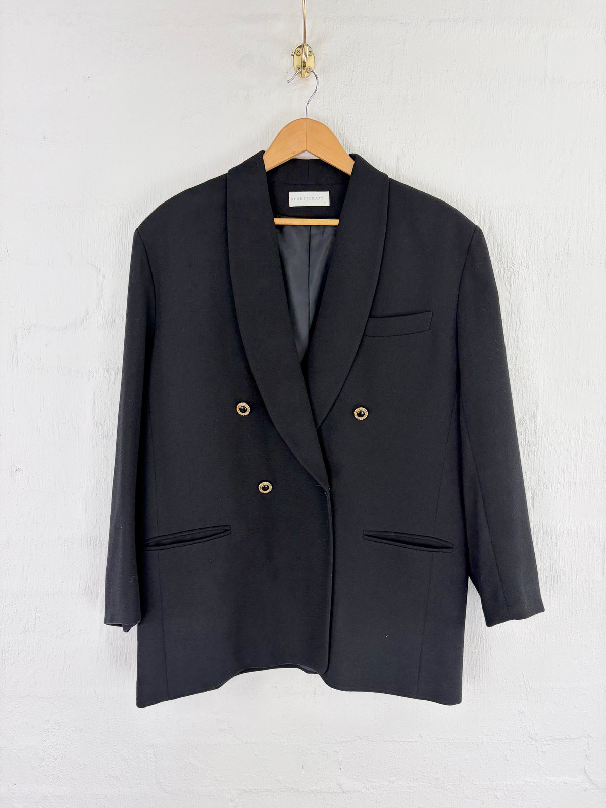 A black double-breasted blazer with shawl lapels and gold-tone buttons, hanging on a wooden hanger against a white painted brick wall.