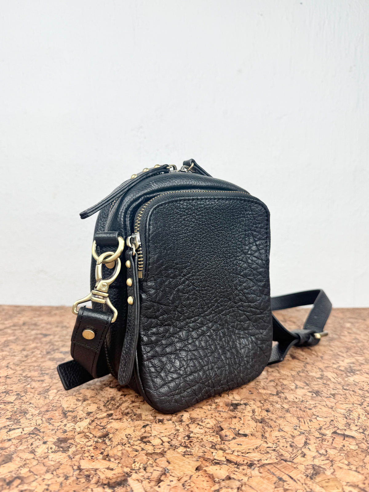 Black pebbled leather crossbody bag with gold-tone hardware, shown on a cork surface against a white wall.