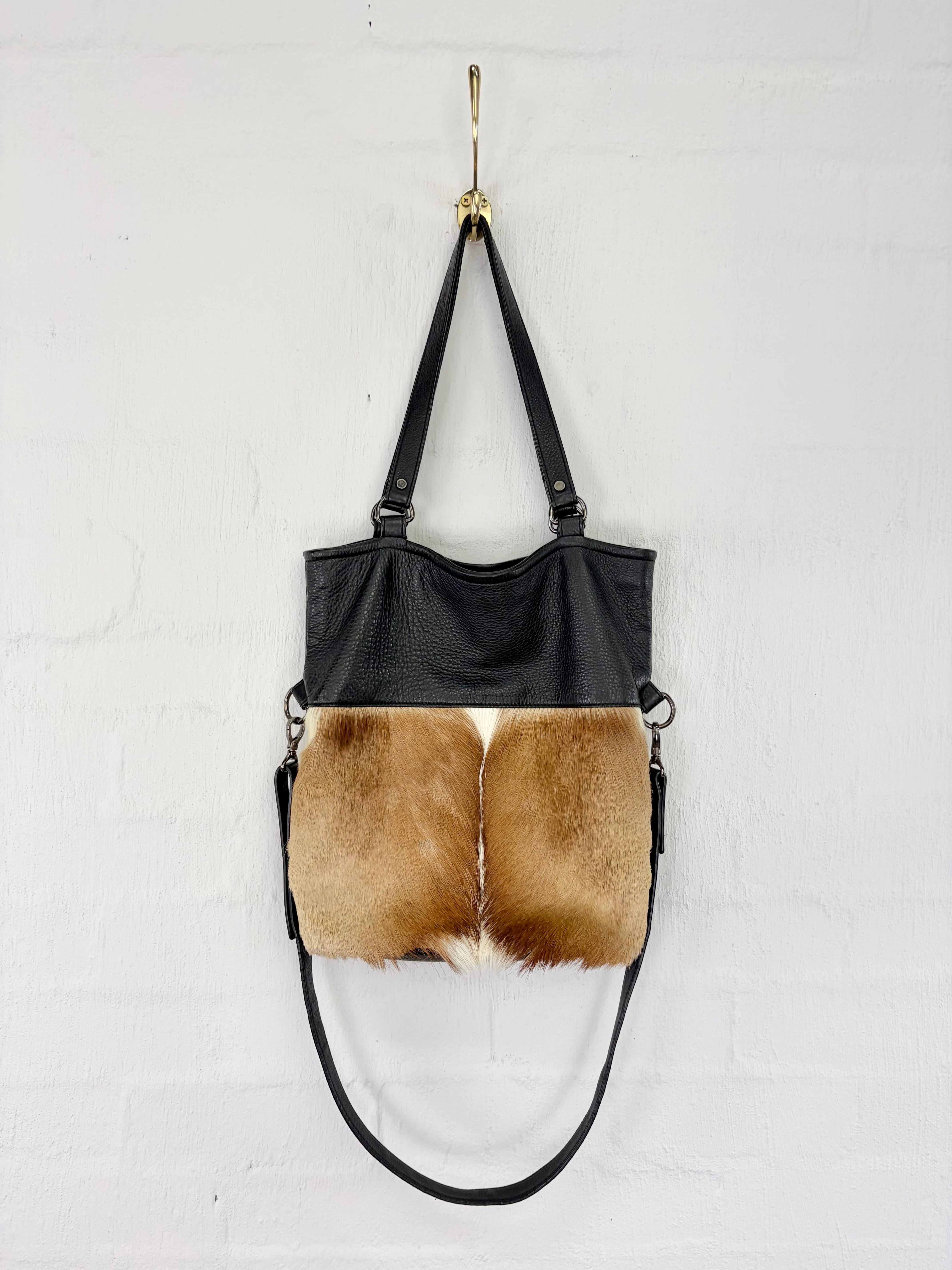 A black faux-leather shoulder bag with a natural tan and white cowhide panel on the front, hanging from a gold hook against a white brick wall.