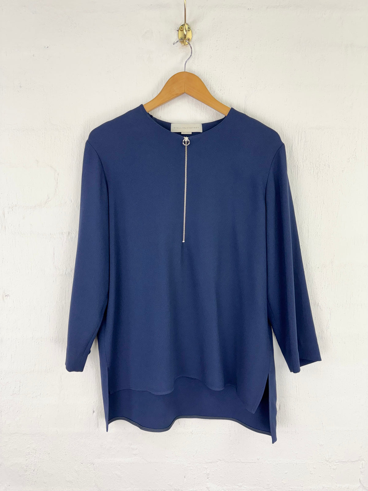 A navy blue long-sleeve top with a silver zipper at the neckline, hanging on a wooden hanger against a white painted brick wall.