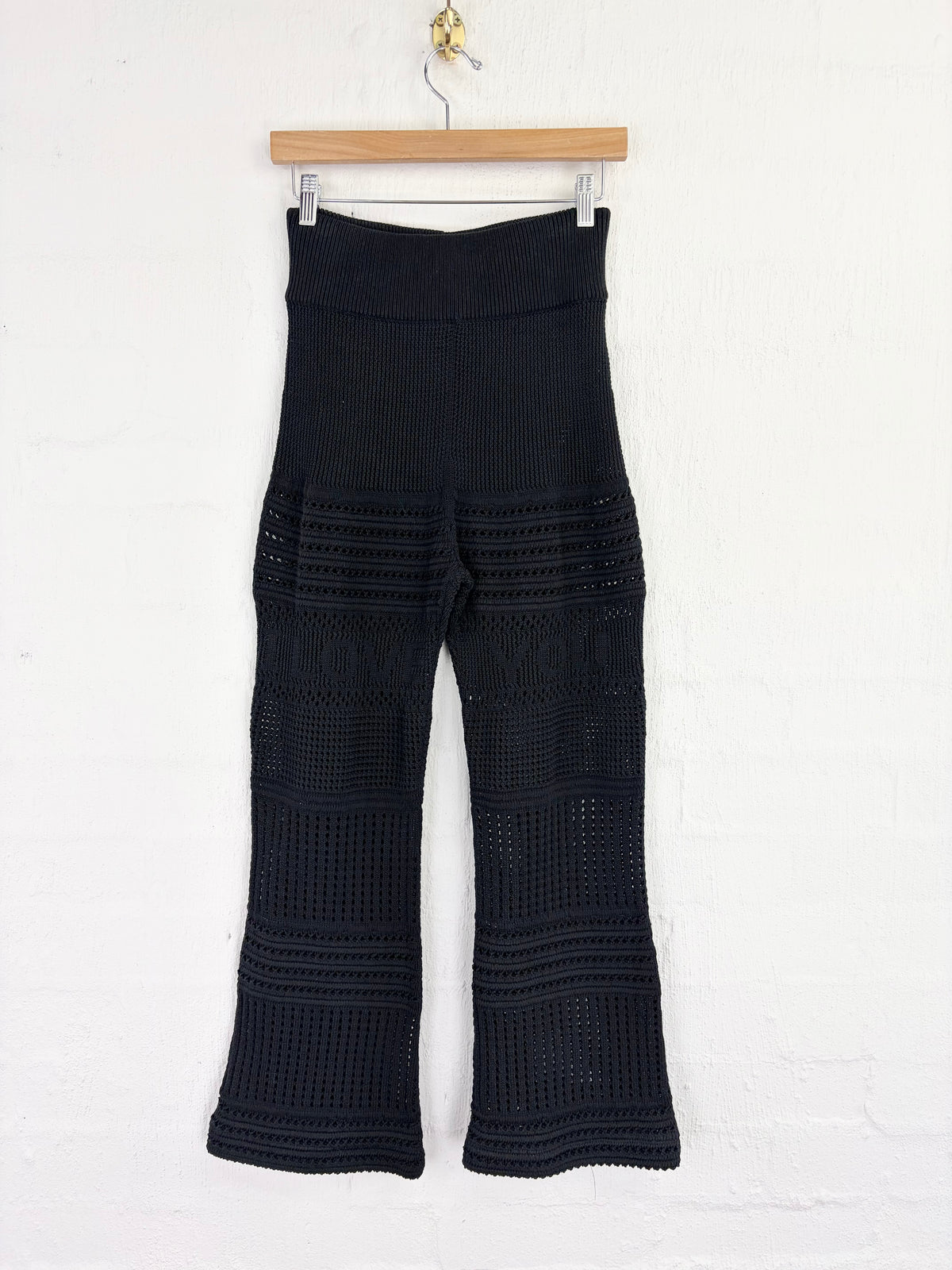 Black crochet knit flare pants hanging from a wooden hanger against a white brick wall.