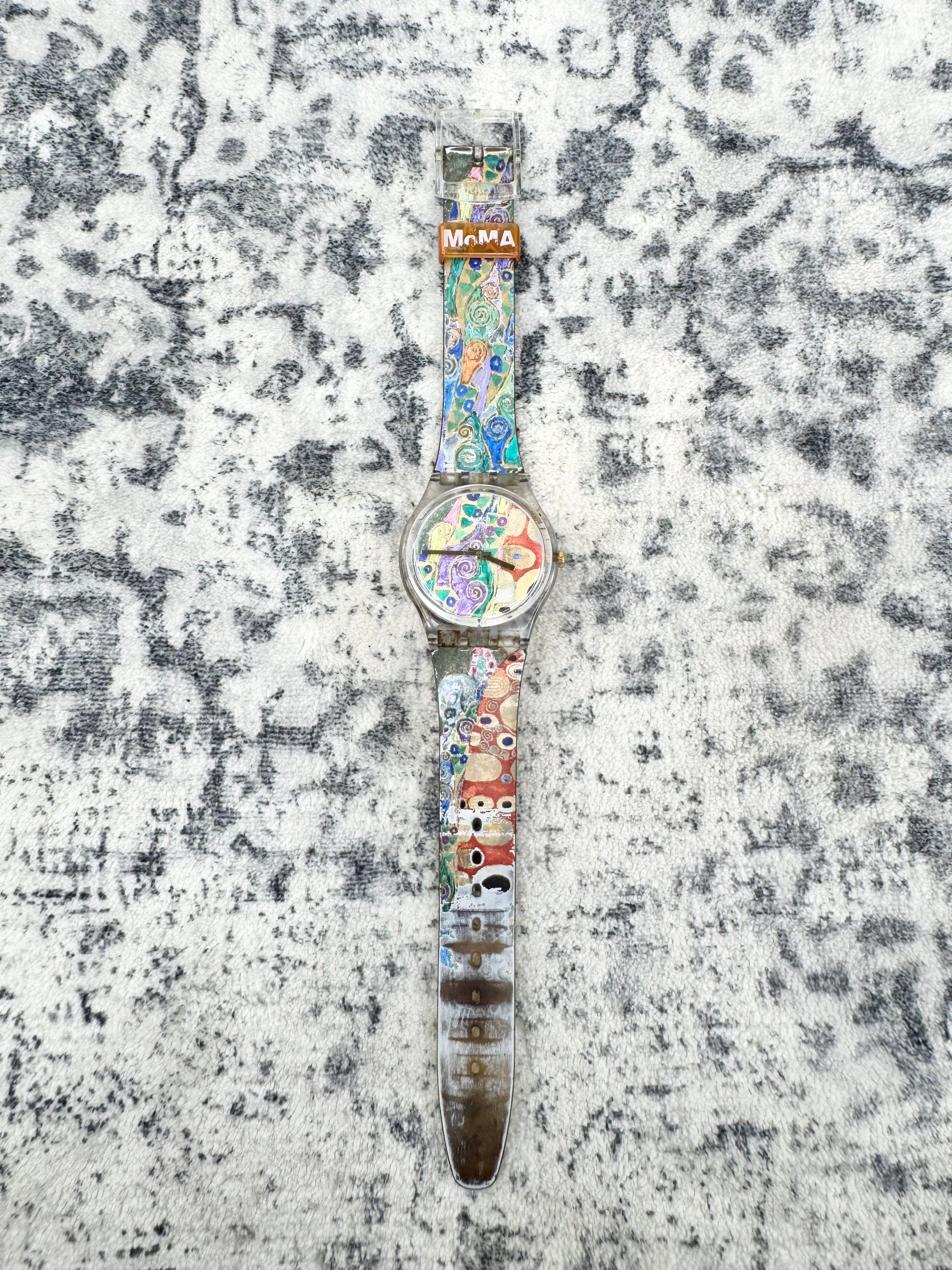 A wristwatch with a colorful, patterned strap and a silver-toned case, displayed flat on a textured gray and white surface. The watch face and strap feature a variety of cartoon-like illustrations, and a small metal tag near the top reads 'MAMA'.
