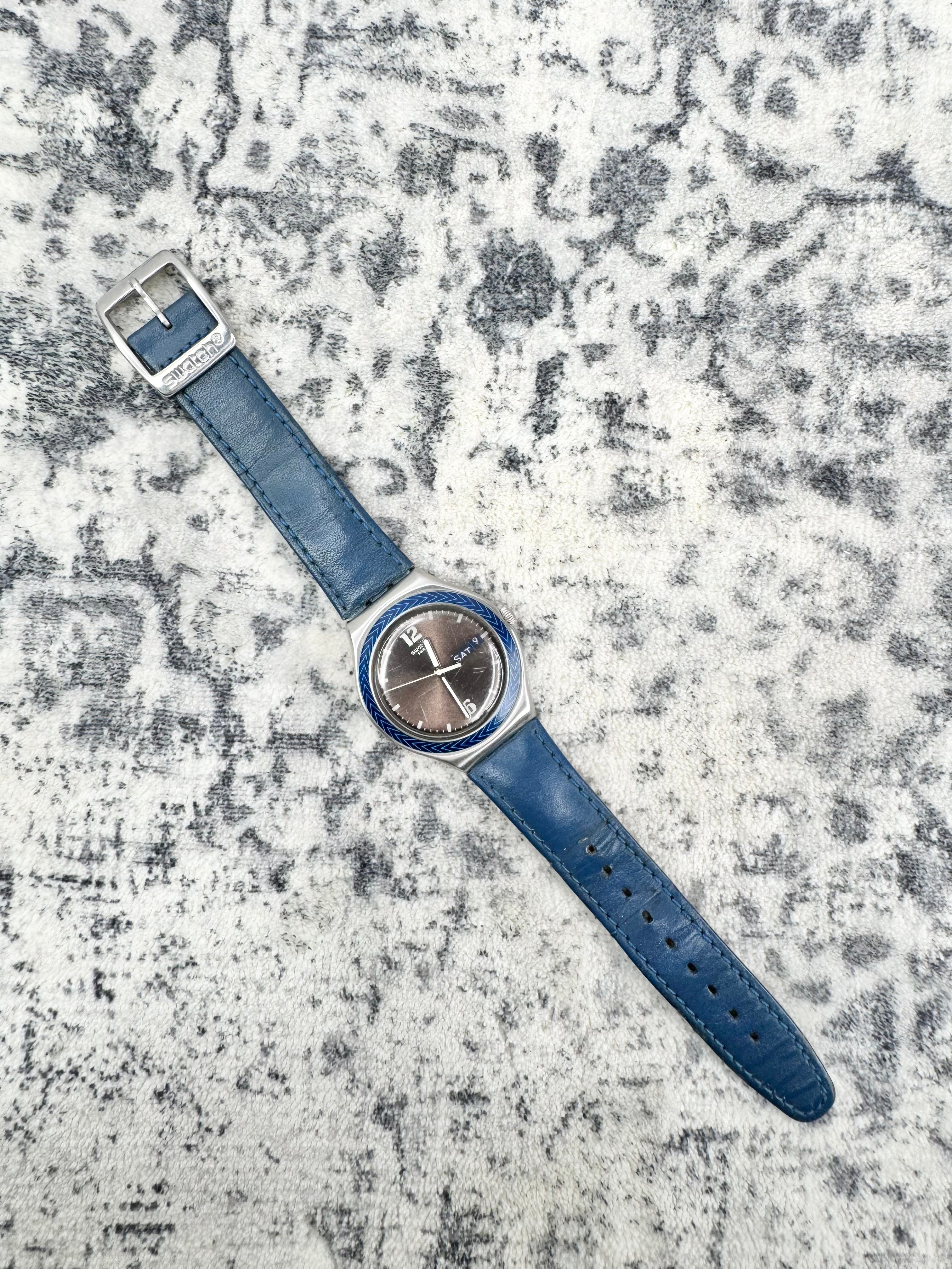 A wristwatch with a blue leather strap, silver-toned case, and a dark gray dial, laid diagonally on a textured gray and white patterned surface.