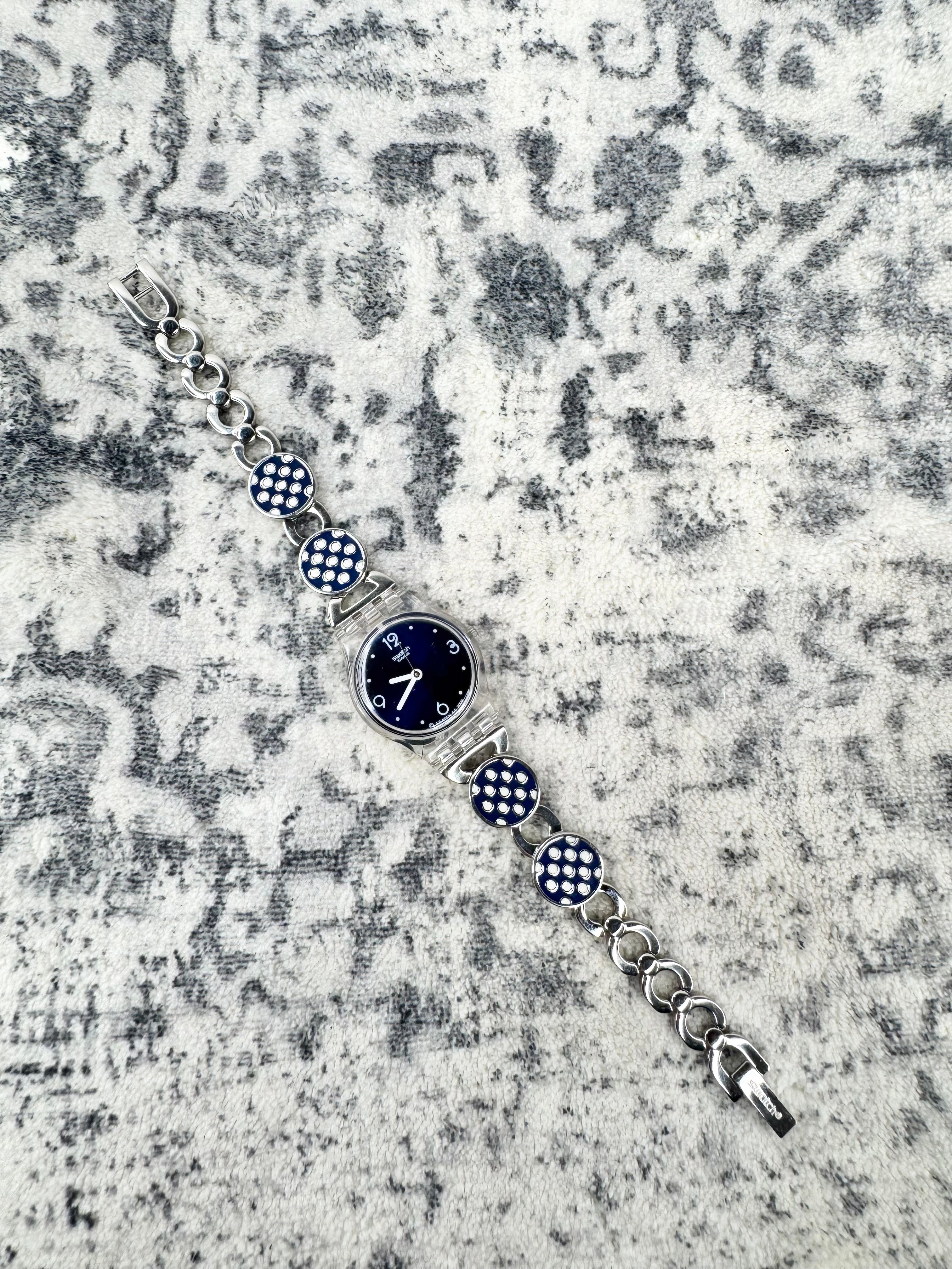 A silver-tone chain watch bracelet featuring a round dark blue dial and three round blue-and-white checkered links, displayed diagonally on a textured gray and white background.