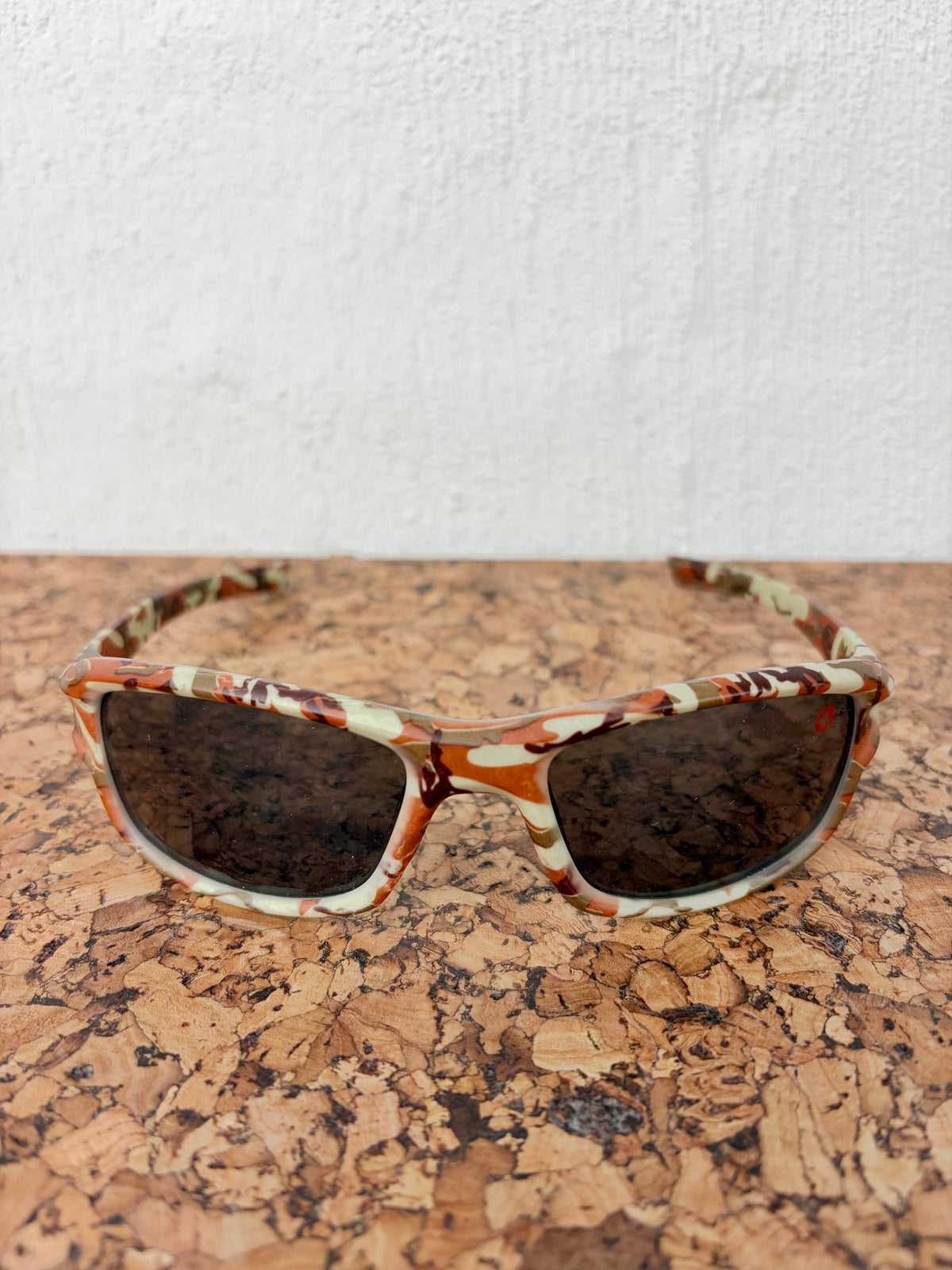 Patterned sunglasses with a brown, orange, and cream abstract design and dark lenses, resting on a cork surface against a white wall.