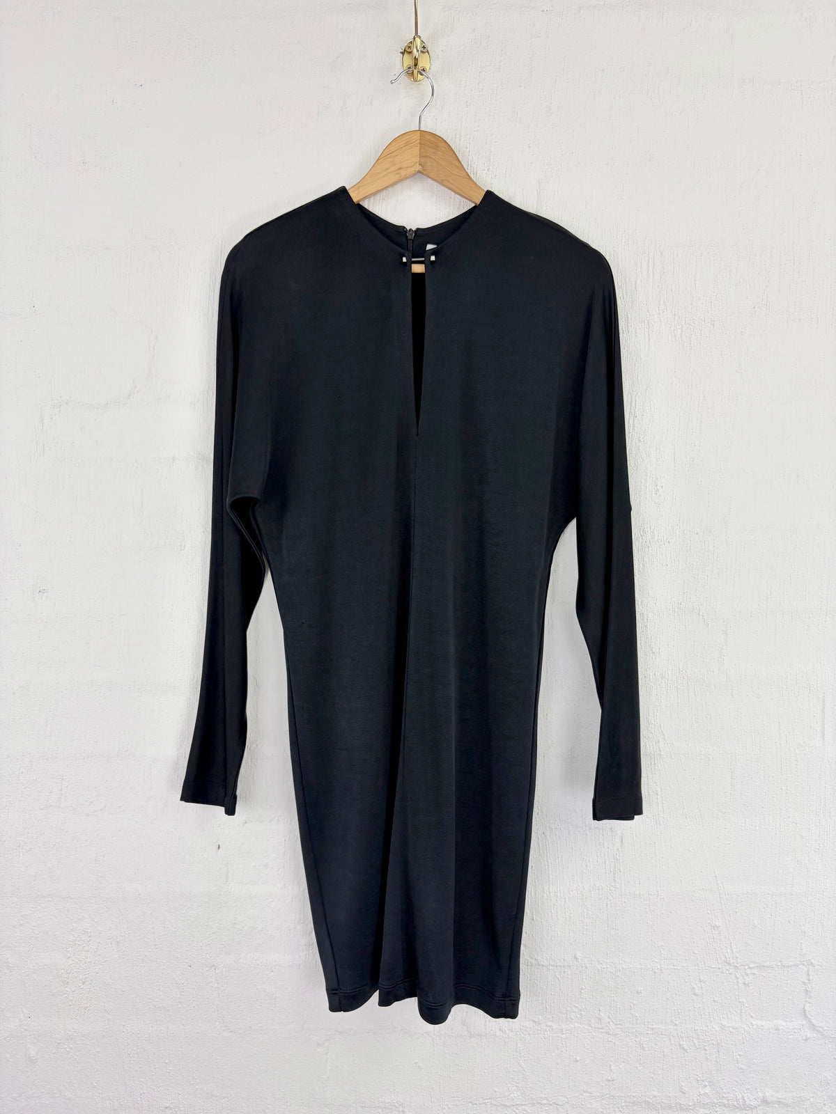 A black long-sleeve dress with a split neckline and front slit, hanging on a wooden hanger against a white brick wall.