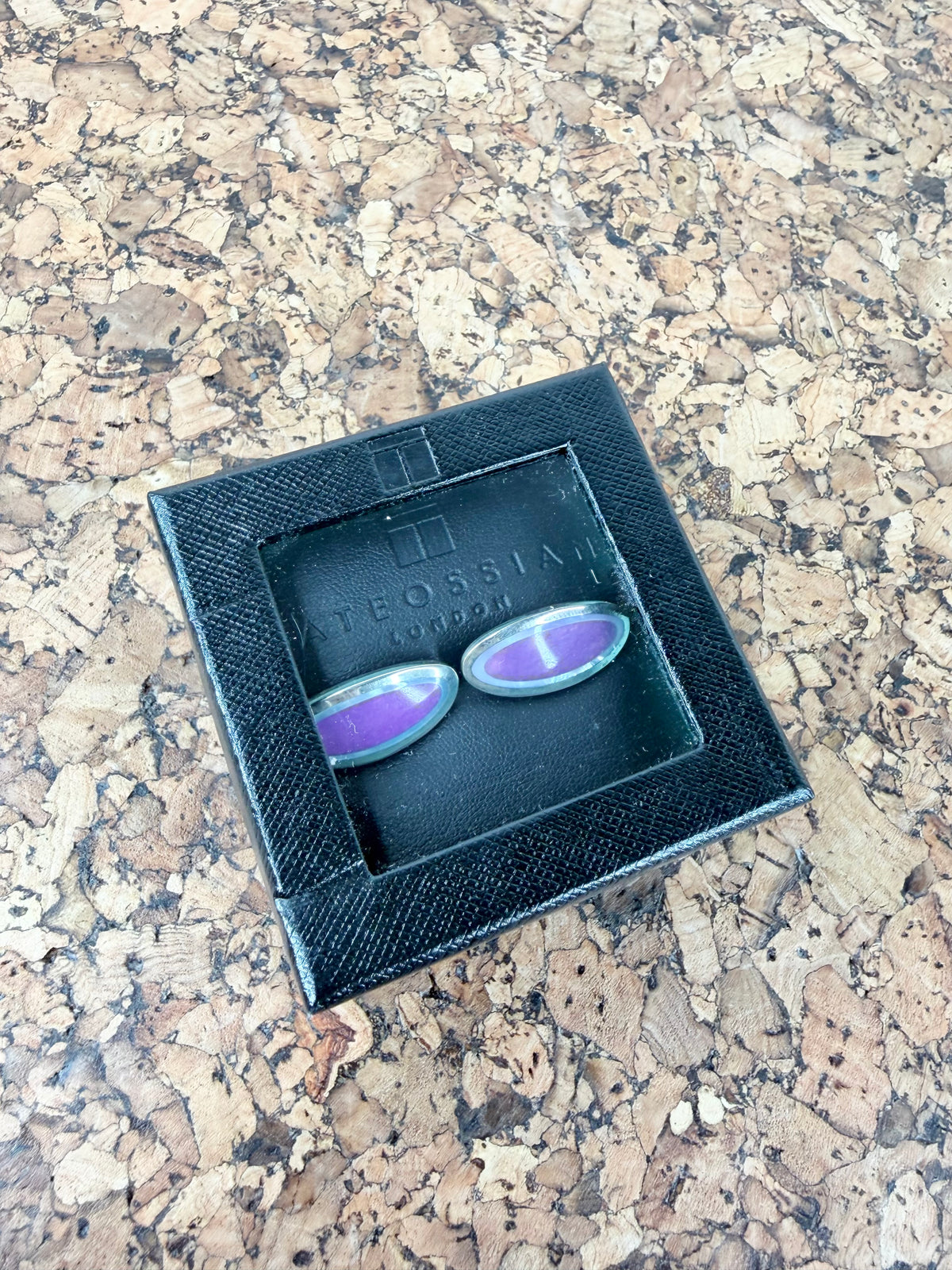 A pair of oval cufflinks with purple inlay and silver-toned metal edges, presented in a black textured box on a cork surface. The brand name 'TEOSSIAN' is embossed inside the box.