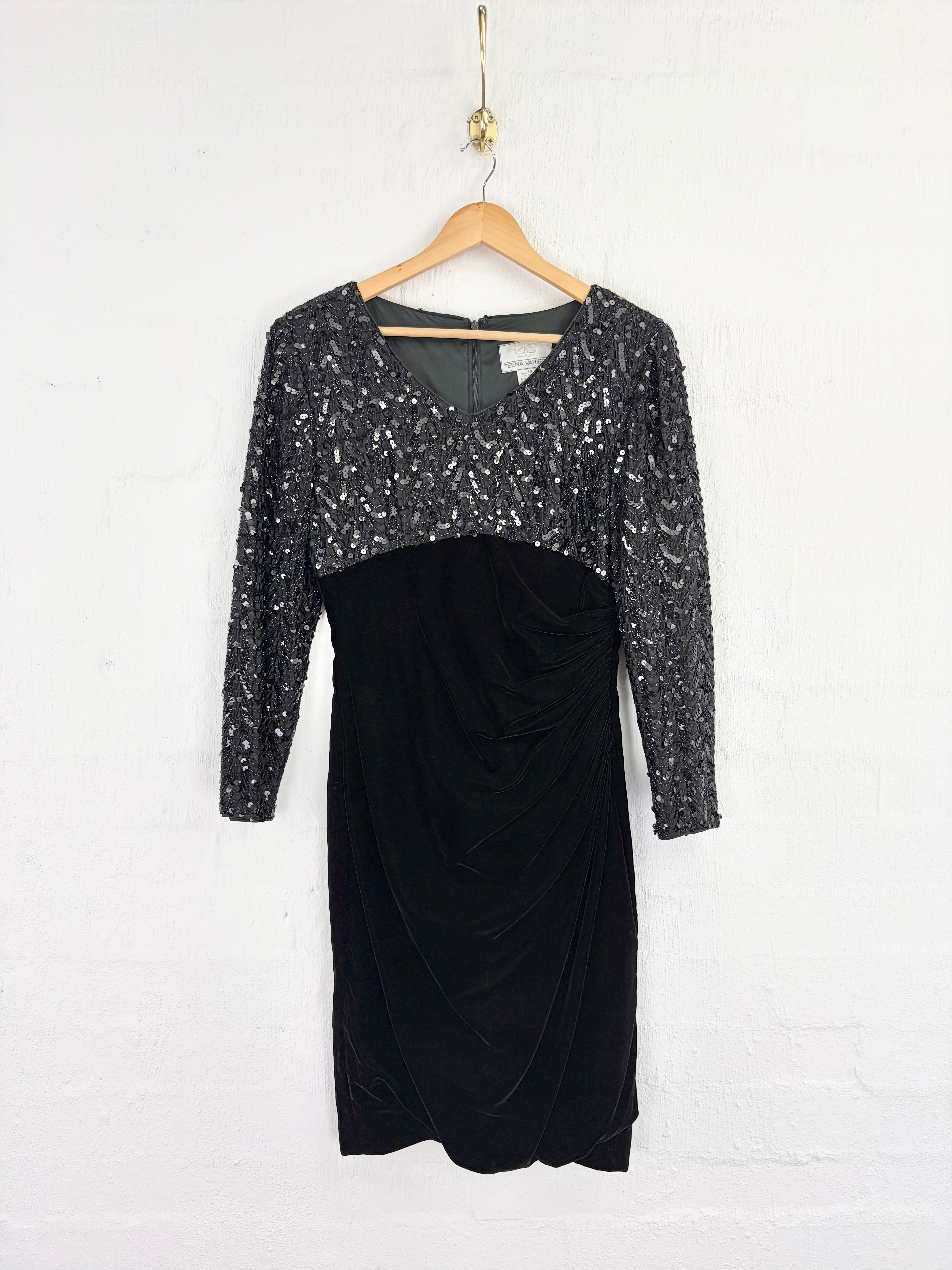 A black long-sleeve dress with a sparkling silver sequined bodice and sleeves, hanging on a wooden hanger against a white brick wall.