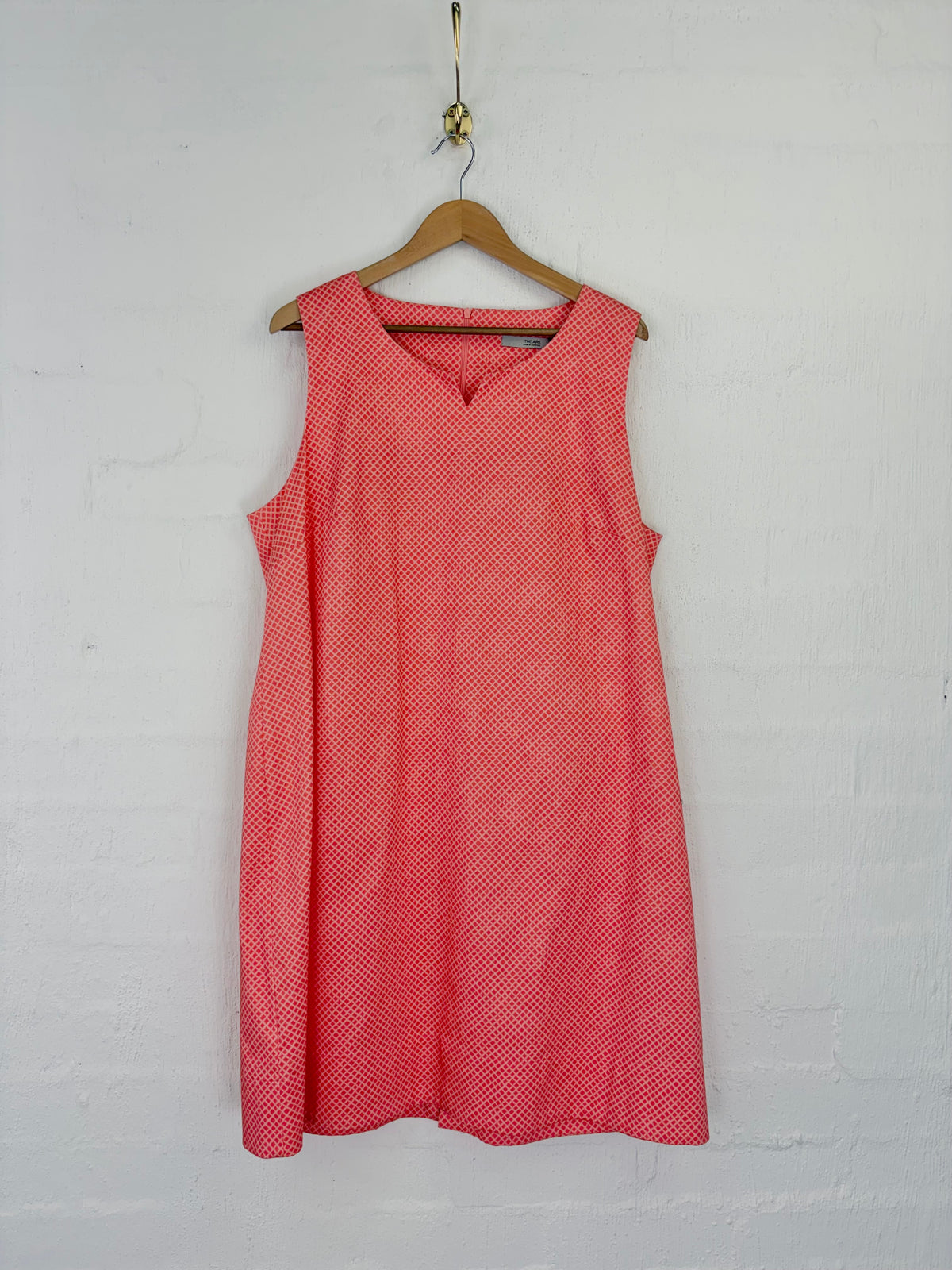 A sleeveless coral pink dress with a small white geometric pattern, hanging on a wooden hanger against a white painted brick wall.
