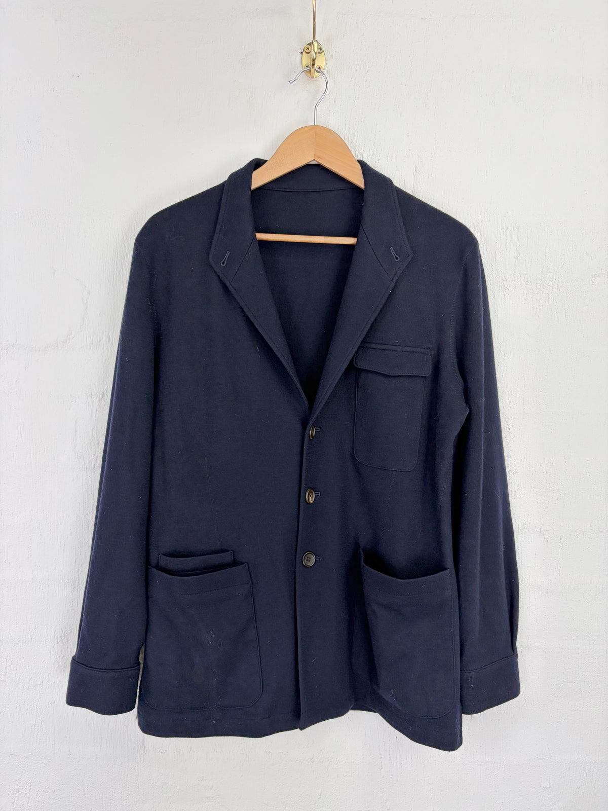 A dark navy blue jacket with a notched lapel, button front, and four front pockets, hanging on a wooden hanger against a white brick wall.