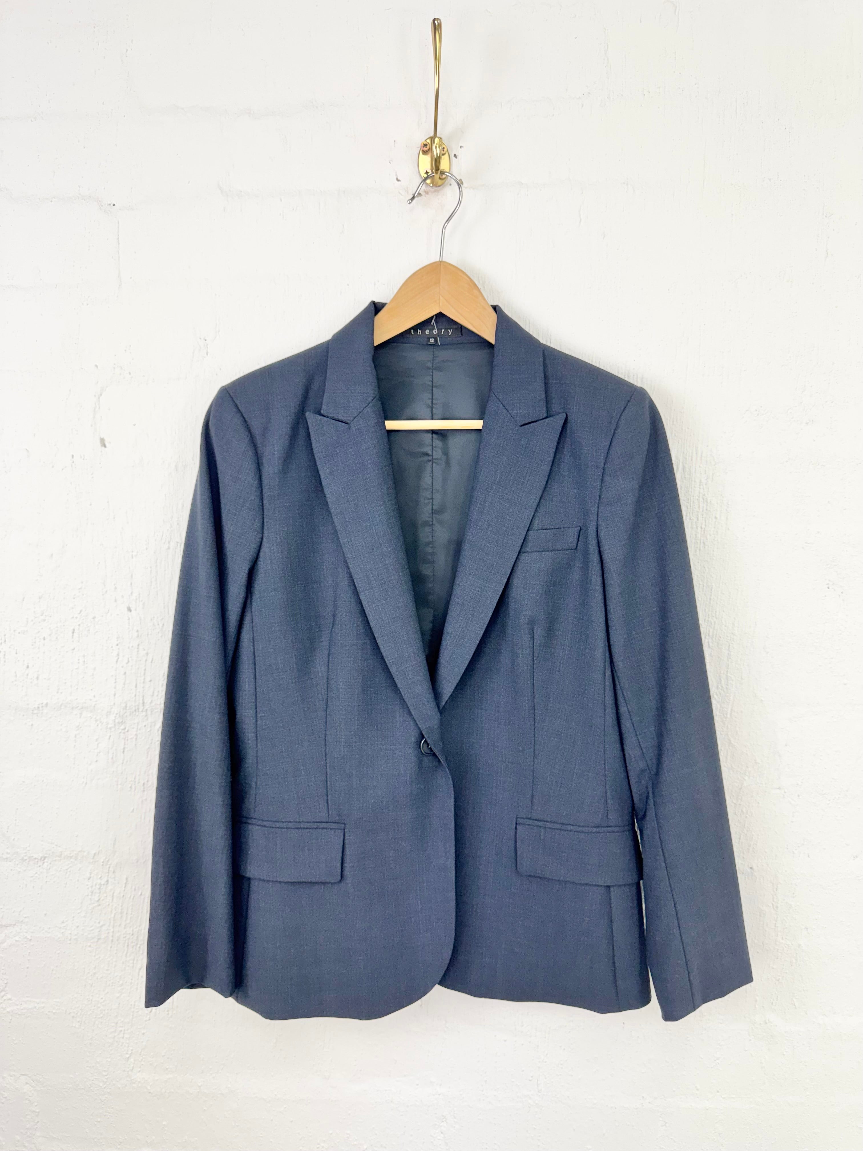 A slate blue single-breasted blazer jacket hanging on a wooden hanger from a gold hook against a white painted brick wall.