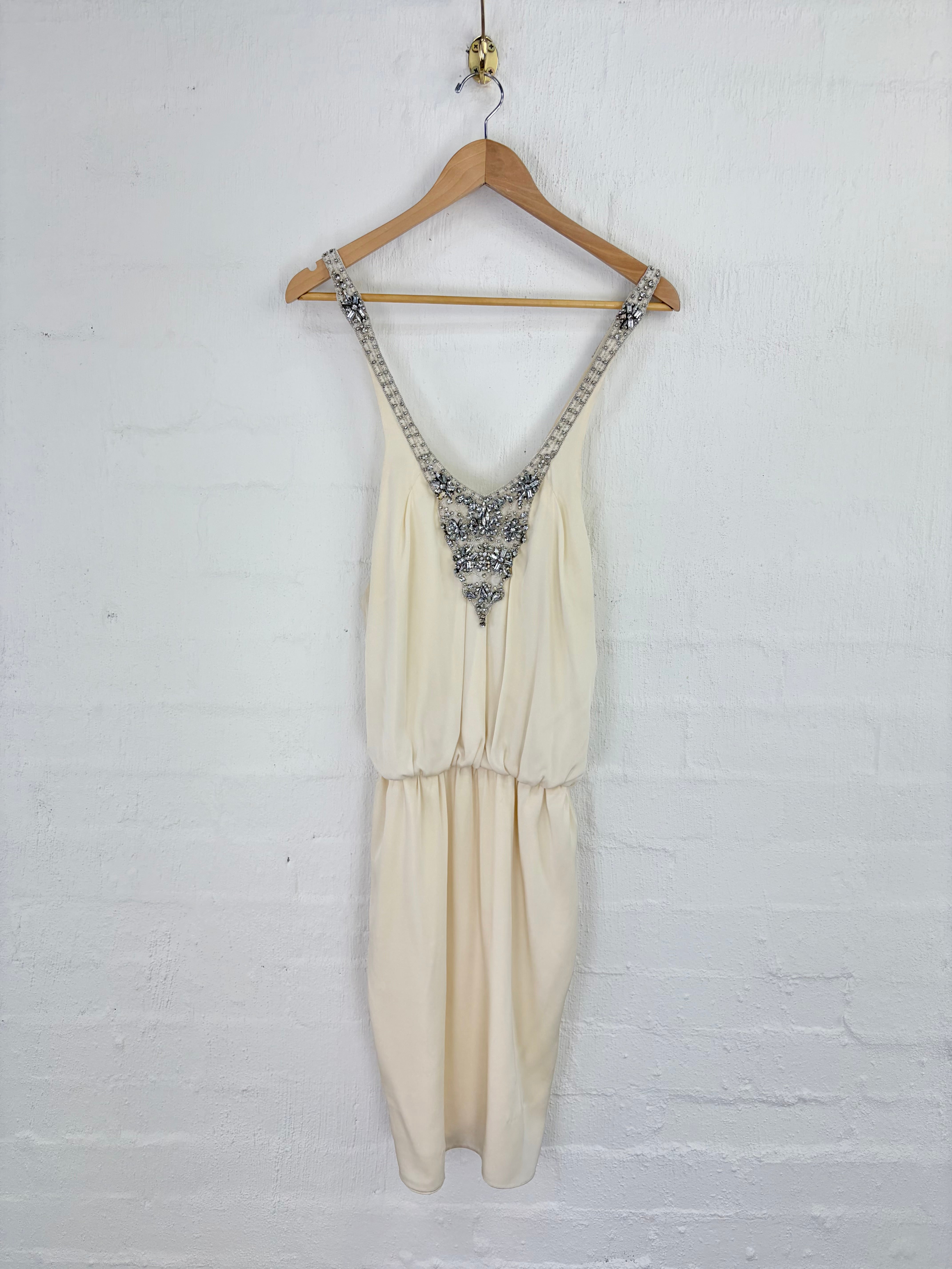 An off-white sleeveless dress with silver beaded straps and a beaded V-neckline, hanging on a wooden hanger against a white brick wall.