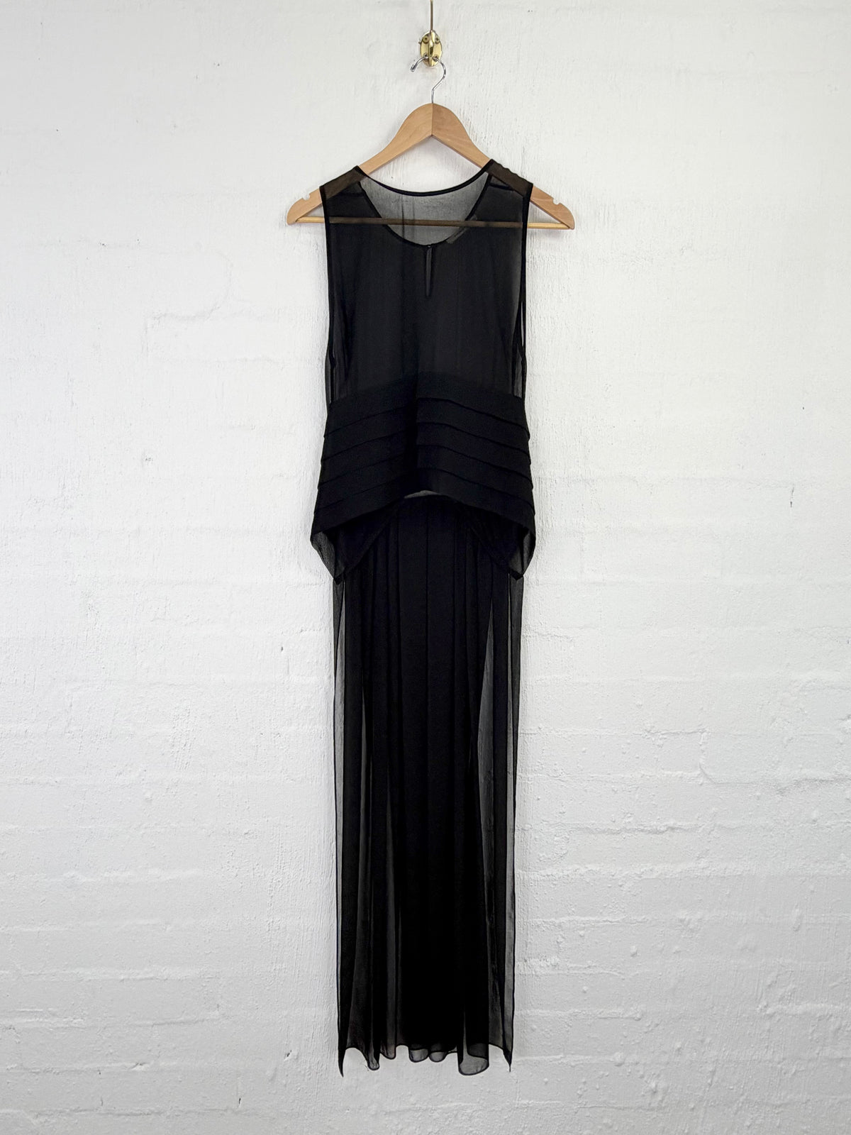 A sleeveless black sheer maxi dress with a tiered waist and flowing pleated skirt, hanging on a wooden hanger against a white painted brick wall.