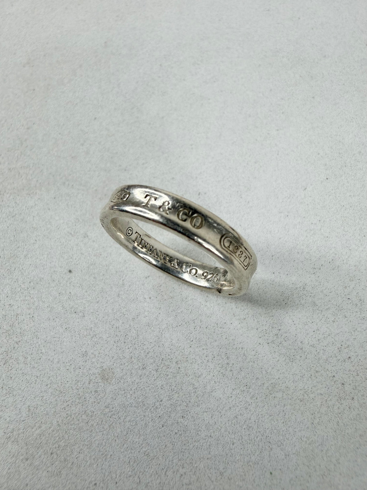 A sterling silver band ring engraved with 'T & CO' and '© THURSDAY 10.27' on a light gray textured background.