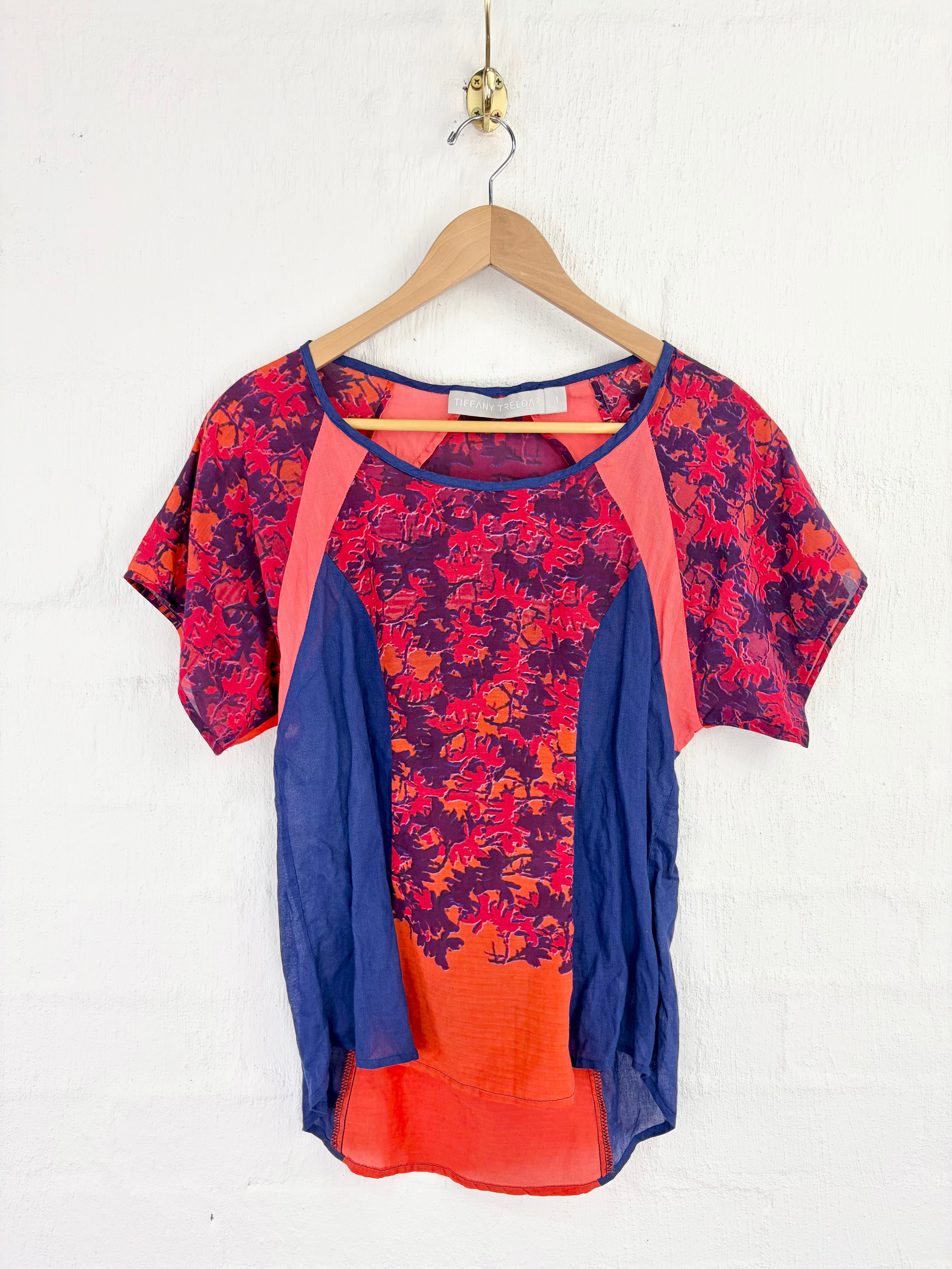 A short-sleeve colorblock top with a red and navy floral pattern, solid coral pink side panels, and solid blue side panels, hanging on a wooden hanger against a white brick wall.