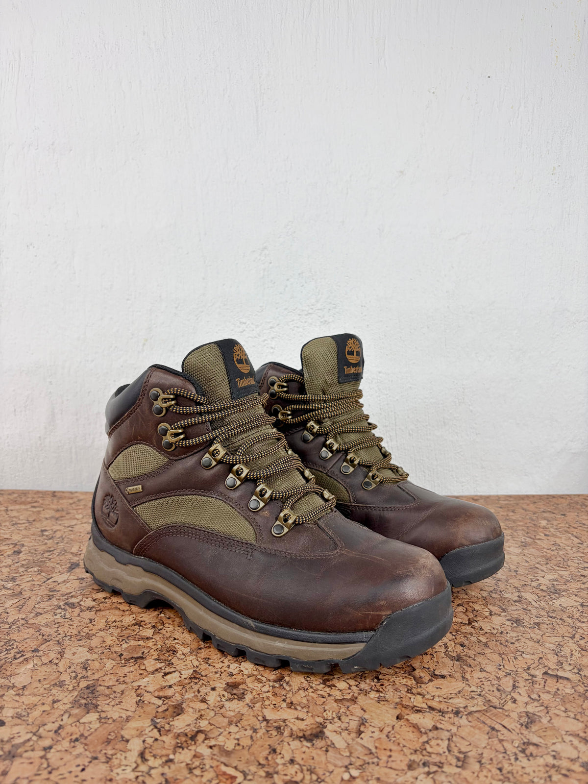 A pair of brown leather hiking boots with olive green accents and laces, displayed on a speckled brown countertop against a white wall.