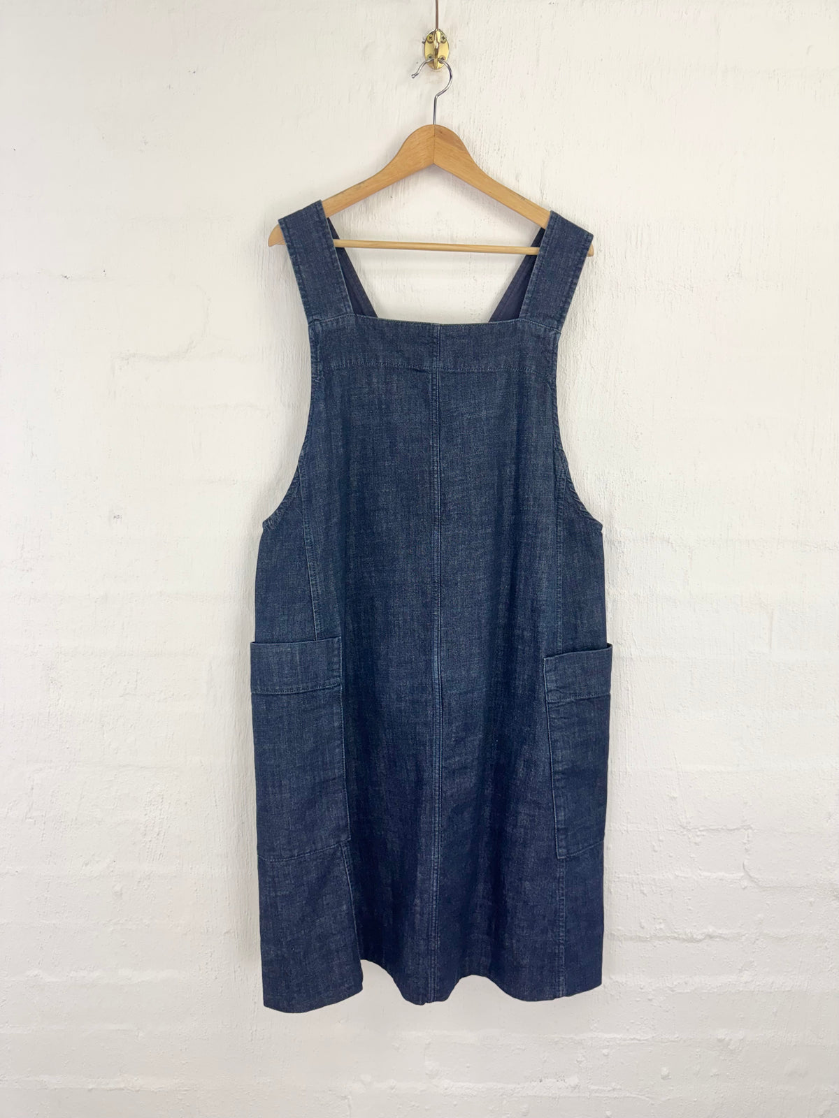A dark blue denim overall dress hanging on a wooden hanger against a white painted brick wall.