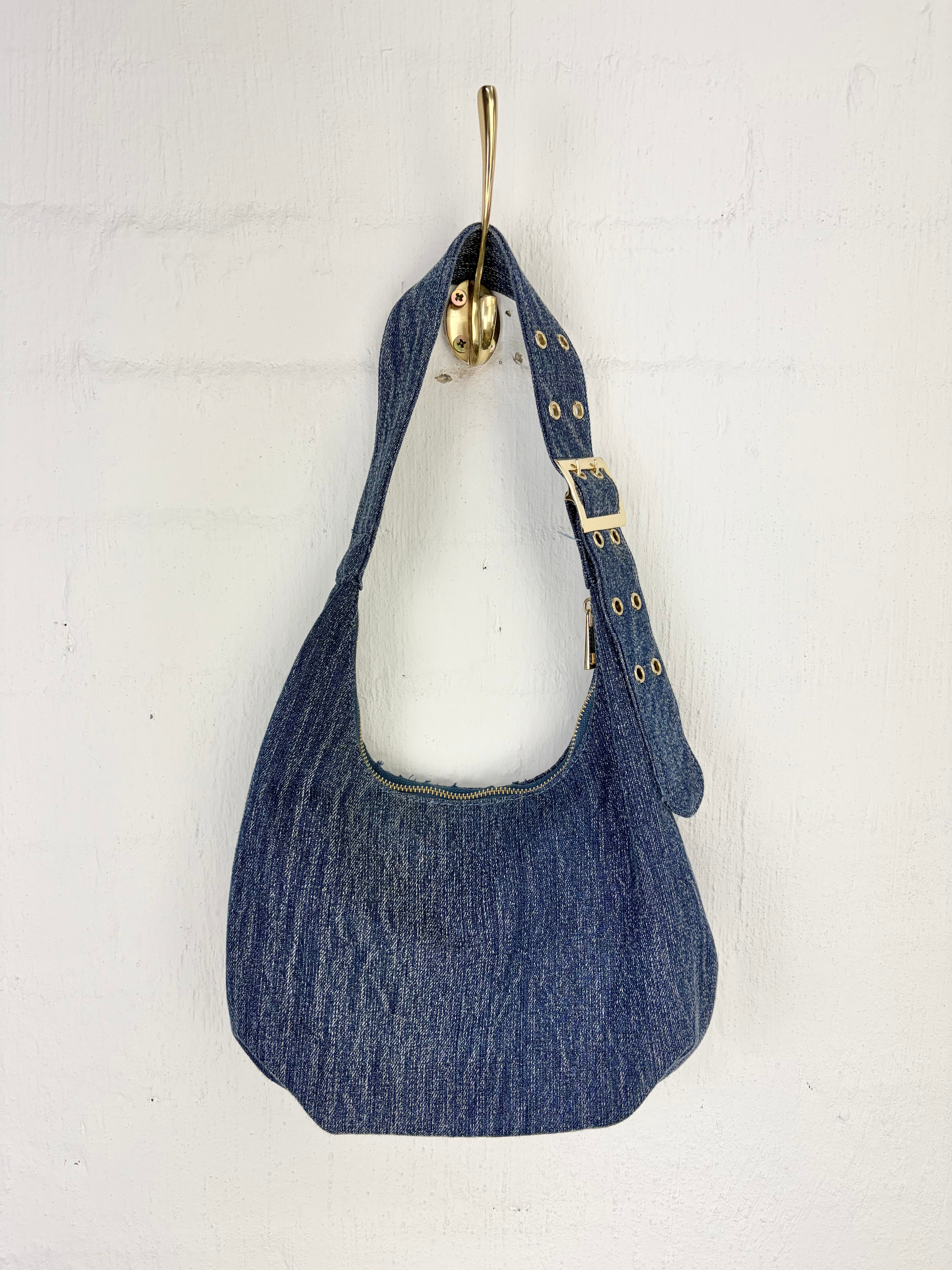 A blue denim shoulder bag with gold-tone hardware, hanging from a gold hook against a white painted brick wall.