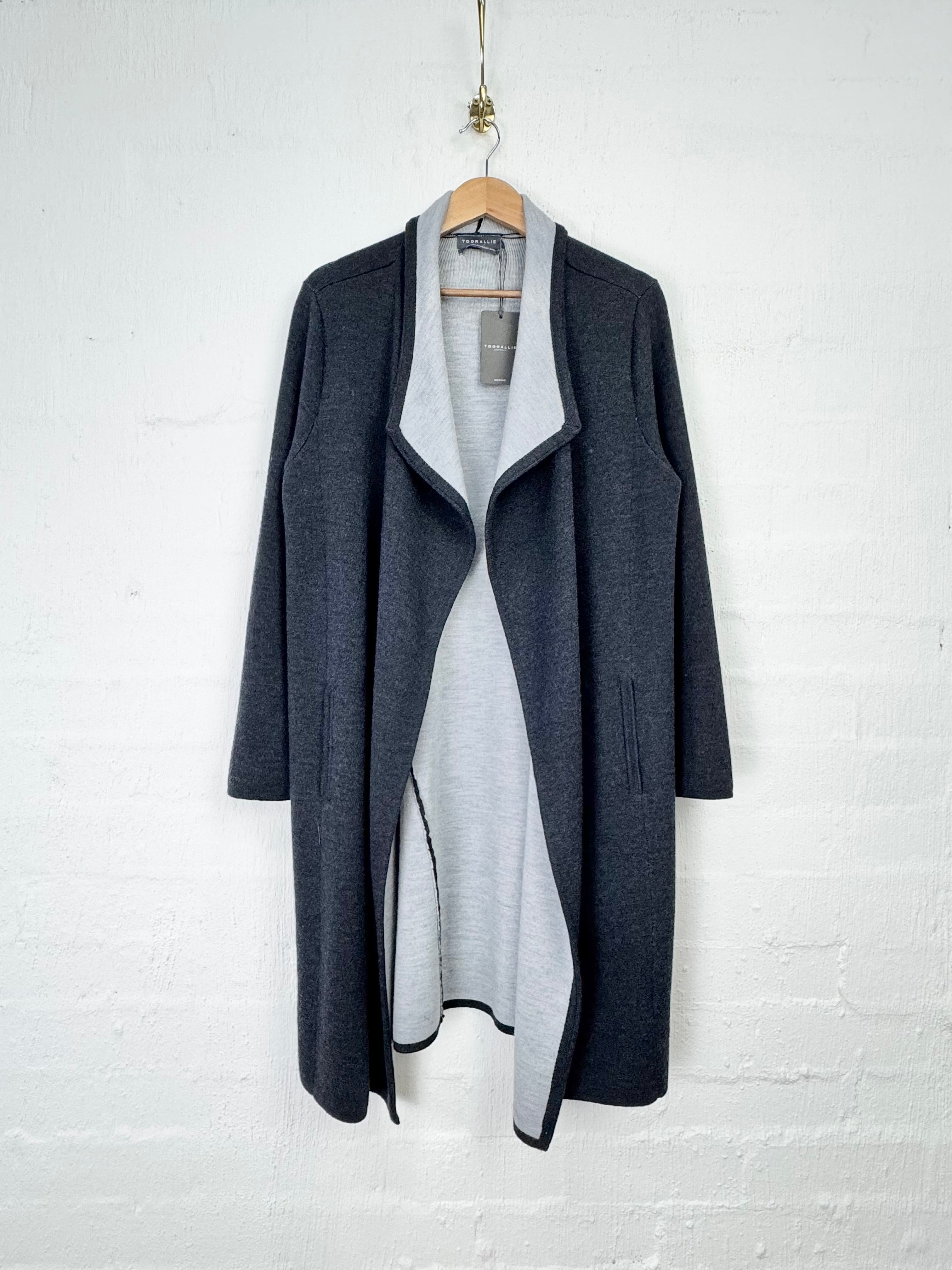A dark gray open-front cardigan coat with a wide collar, hanging on a wooden hanger against a white painted brick wall. A light gray knit top is layered underneath.