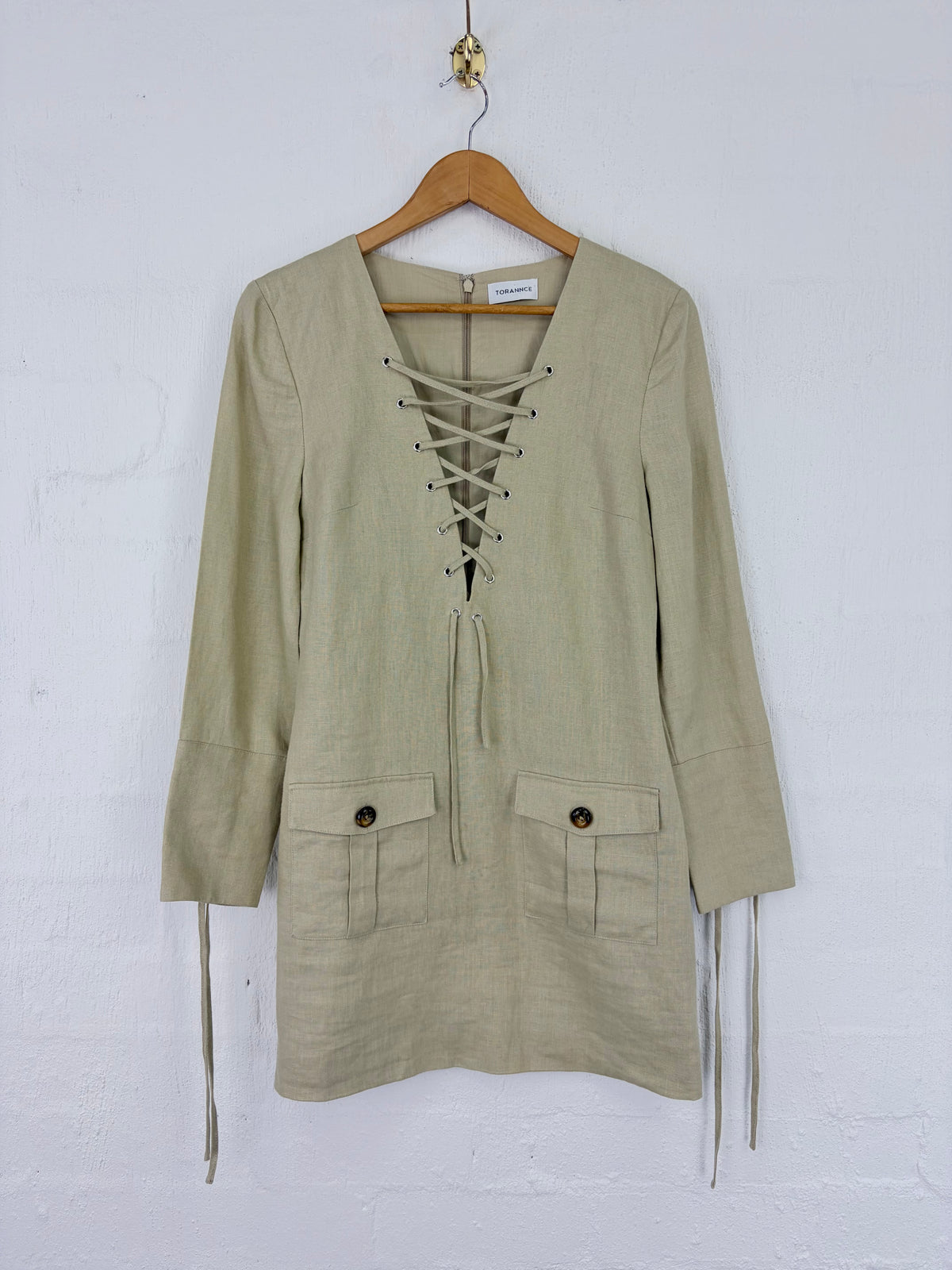 A light beige linen long-sleeve tunic top with a lace-up front, hanging on a wooden hanger against a white painted brick wall.