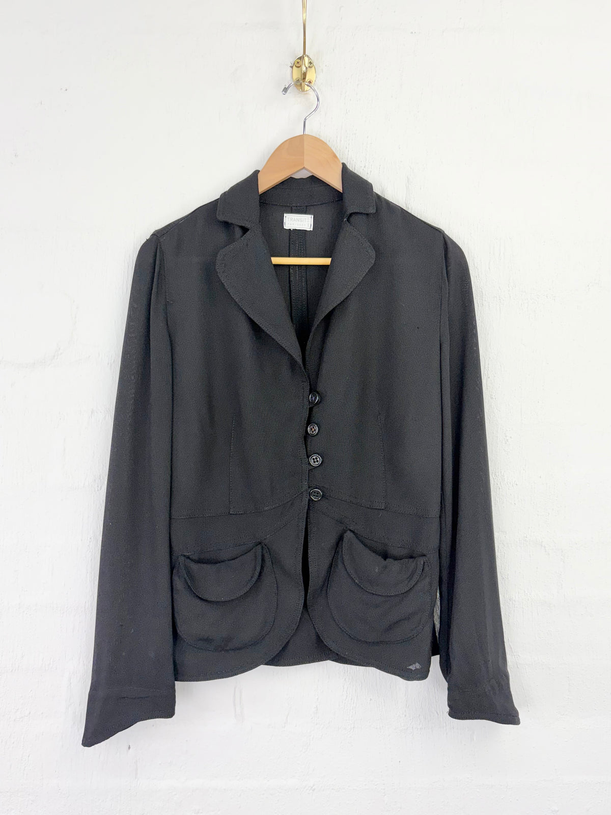 A dark charcoal gray blazer jacket with notch lapels, a three-button front closure, and two large front patch pockets, hanging on a wooden hanger against a white painted brick wall.