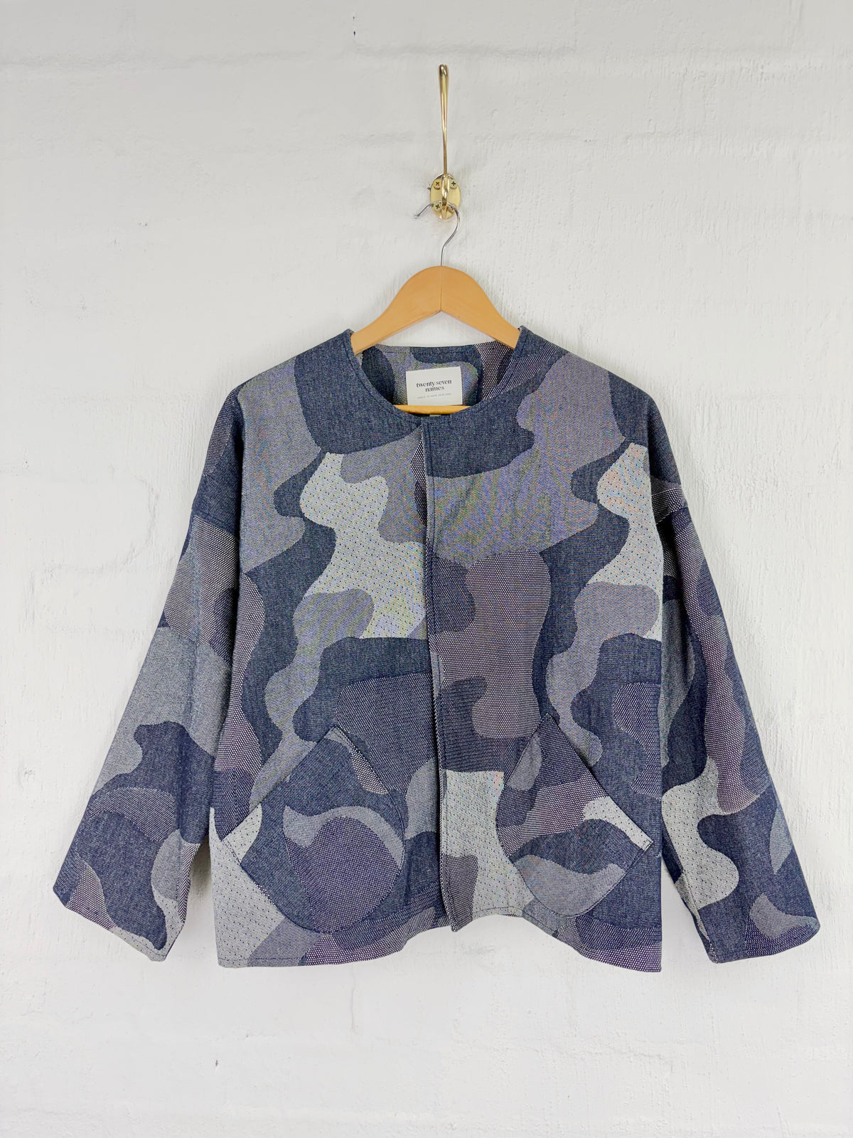 A long-sleeve cropped jacket with an abstract camouflage pattern in shades of slate blue, gray, and lavender, hanging on a wooden hanger against a white brick wall.