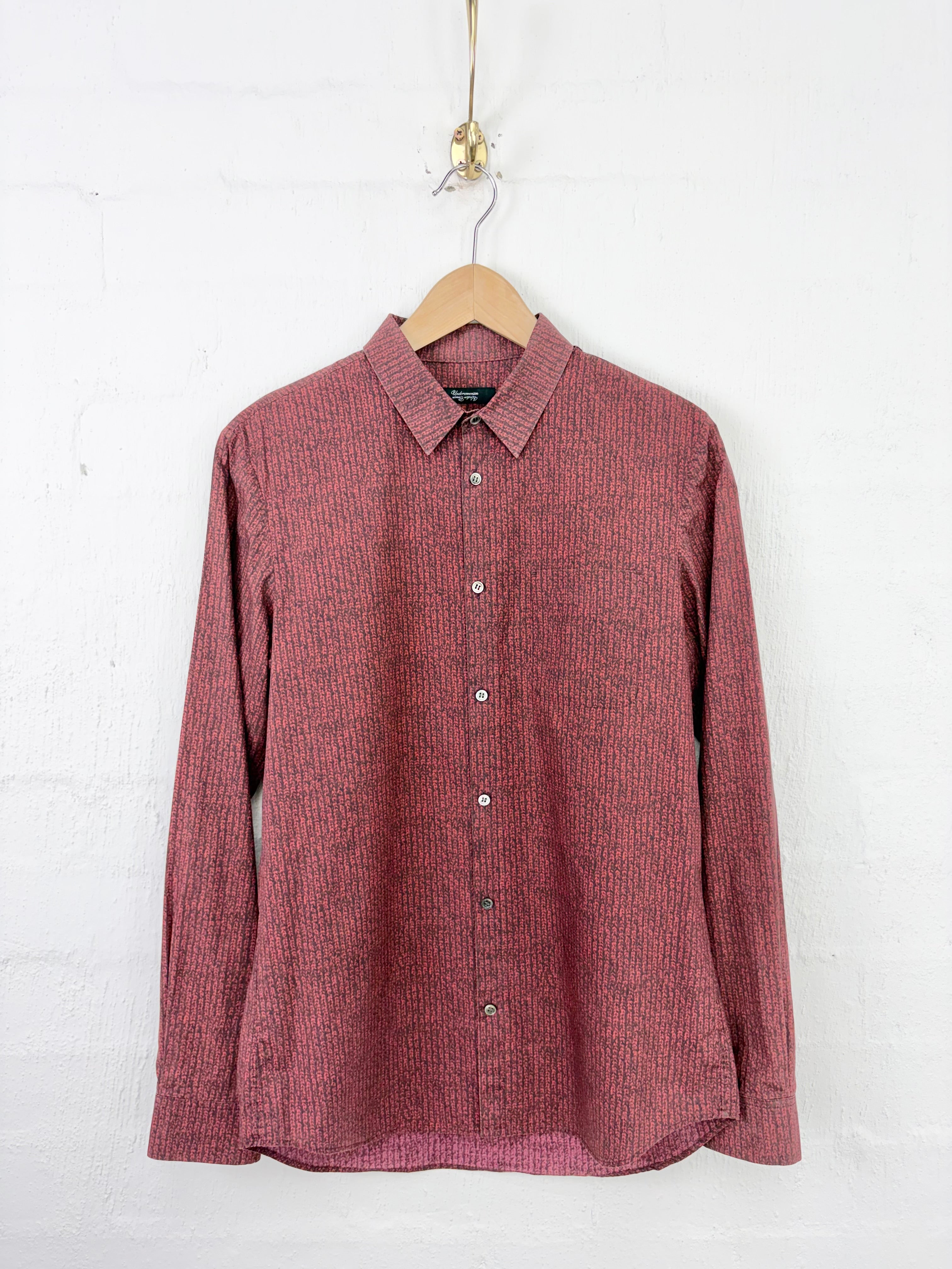 A long-sleeve button-up shirt in a textured, heathered dusty rose or muted red fabric, hanging on a wooden hanger against a white painted brick wall.