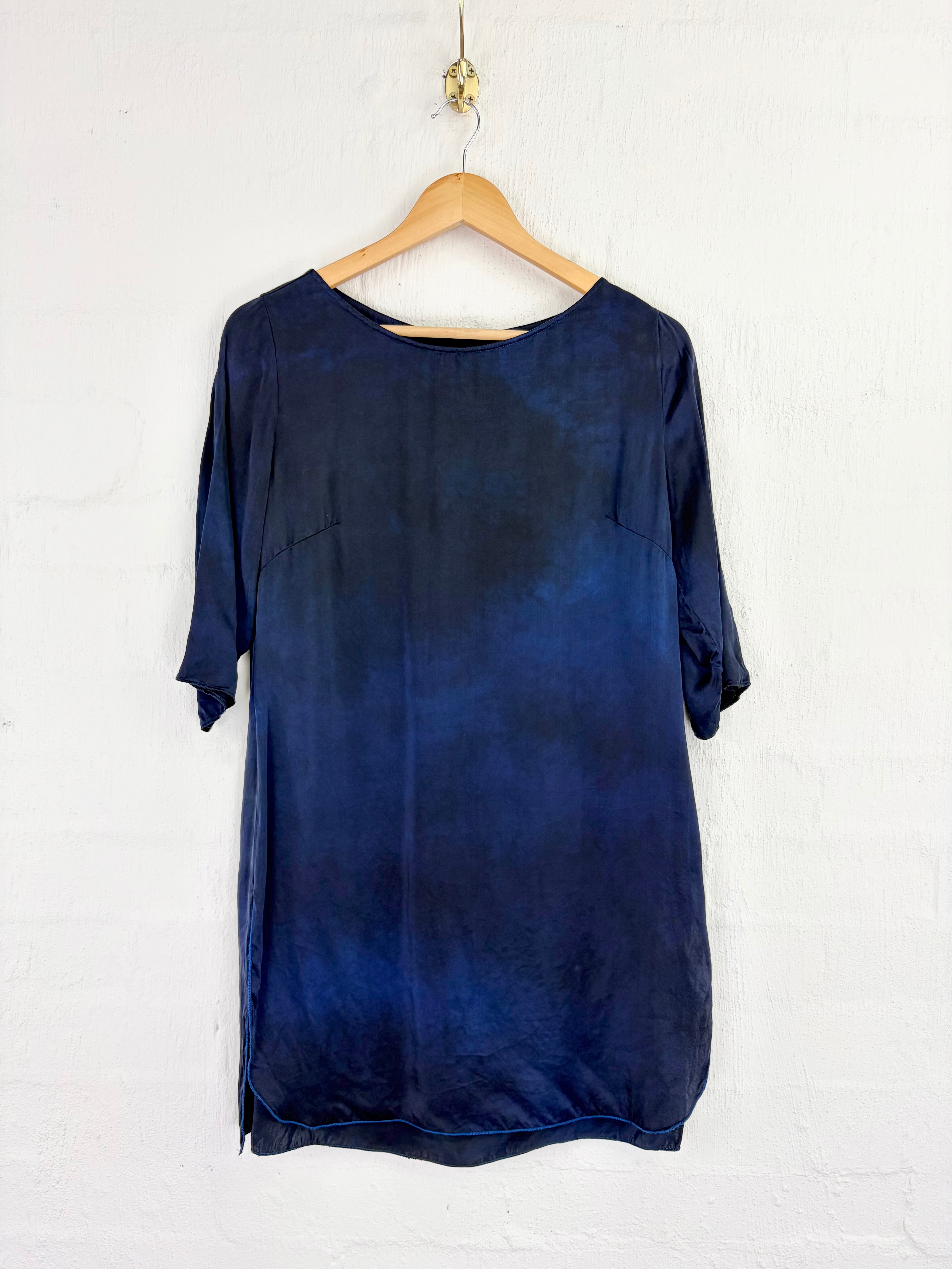 A dark blue tie-dye tunic top with three-quarter sleeves hanging on a wooden hanger against a white painted brick wall.