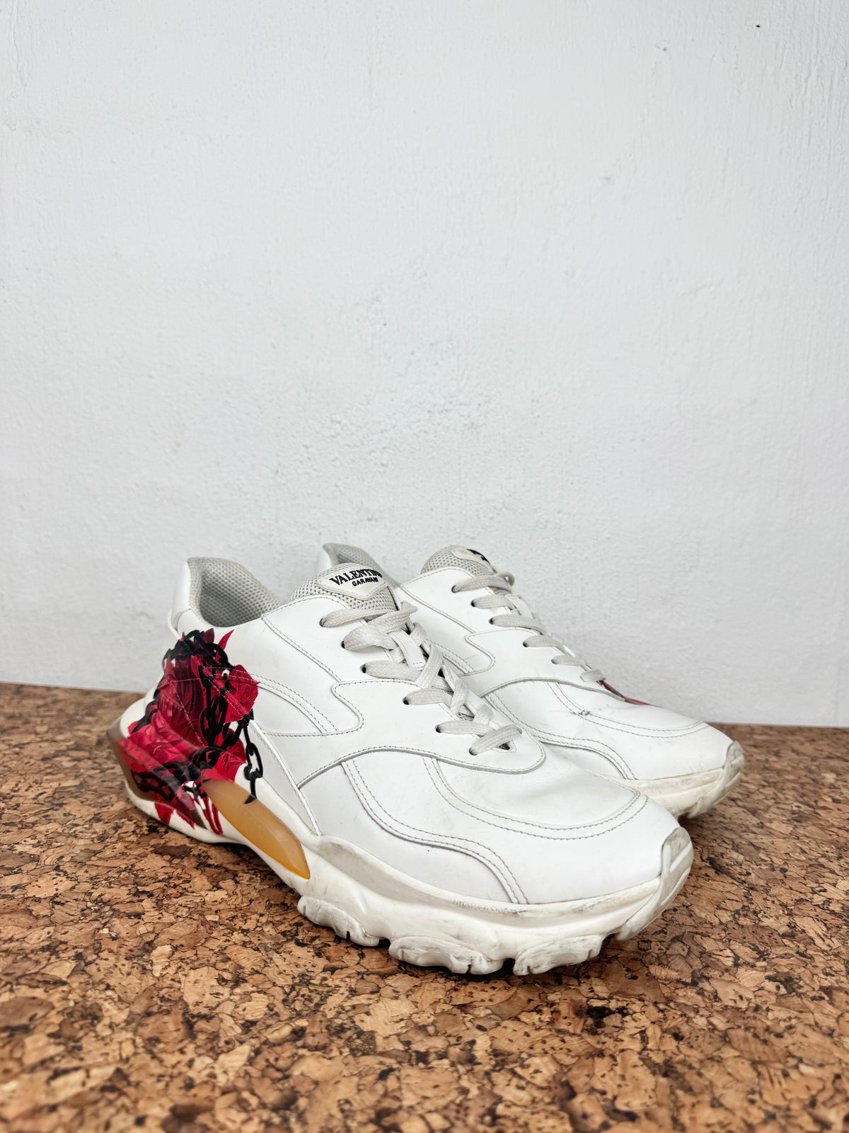 A pair of white leather sneakers with chunky white soles, featuring a prominent red rose graphic on the heel of the front shoe and a gold accent panel on the midsole.