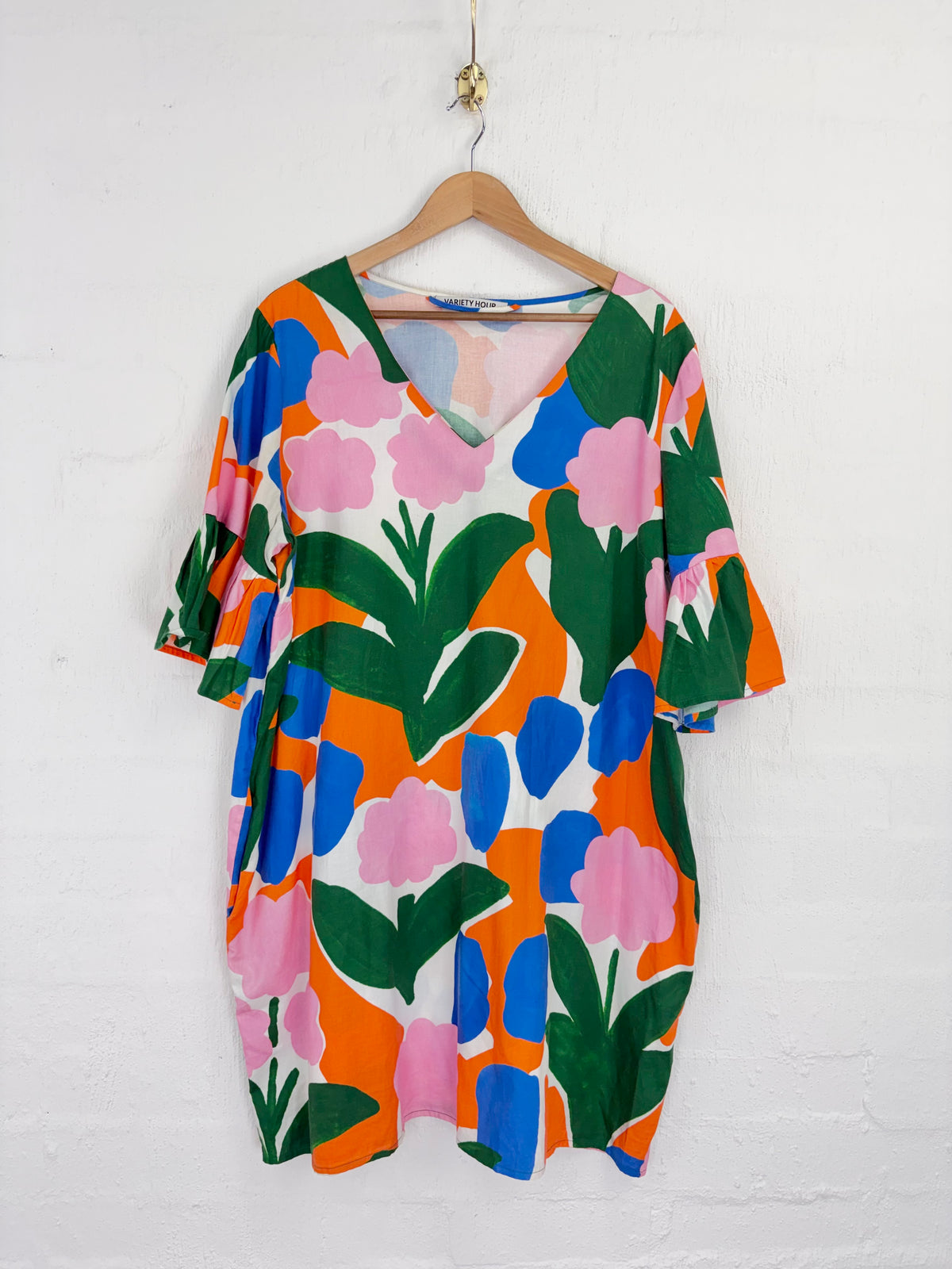 A short-sleeve V-neck dress with a bold, colorful abstract floral pattern in orange, pink, blue, and green, hanging on a wooden hanger against a white painted brick wall.
