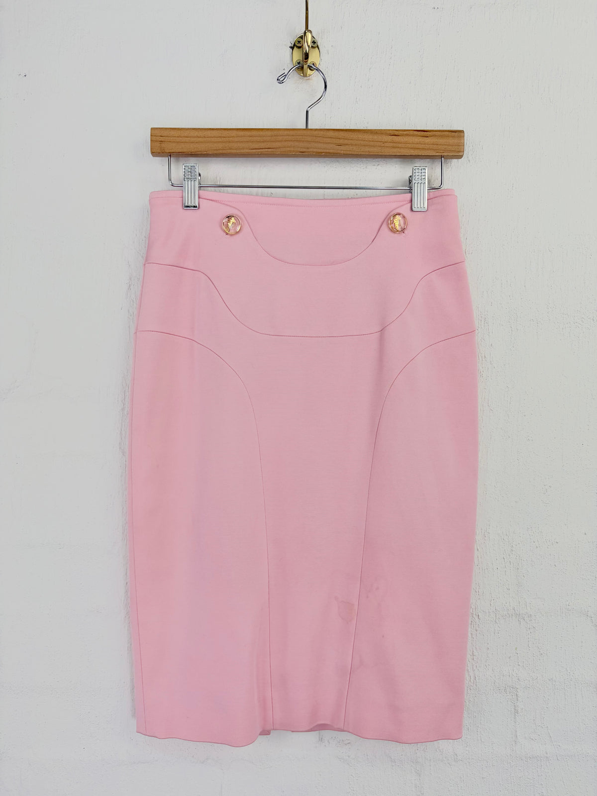 A light pink pencil skirt with structured seams and two gold-tone decorative buttons at the waistband, hanging from a wooden hanger against a white wall.