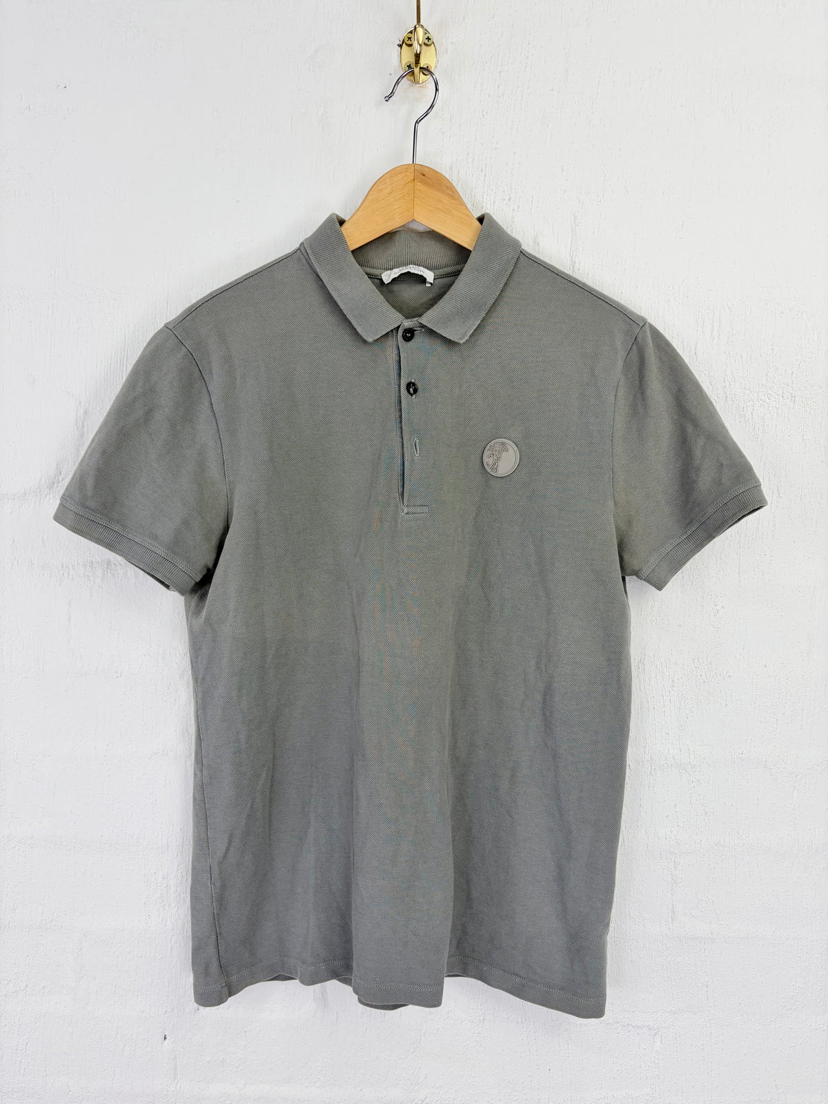 A gray short-sleeve polo shirt hanging on a wooden hanger against a white brick wall. The shirt features a classic collar, a three-button placket, and a small circular metallic emblem on the left chest.