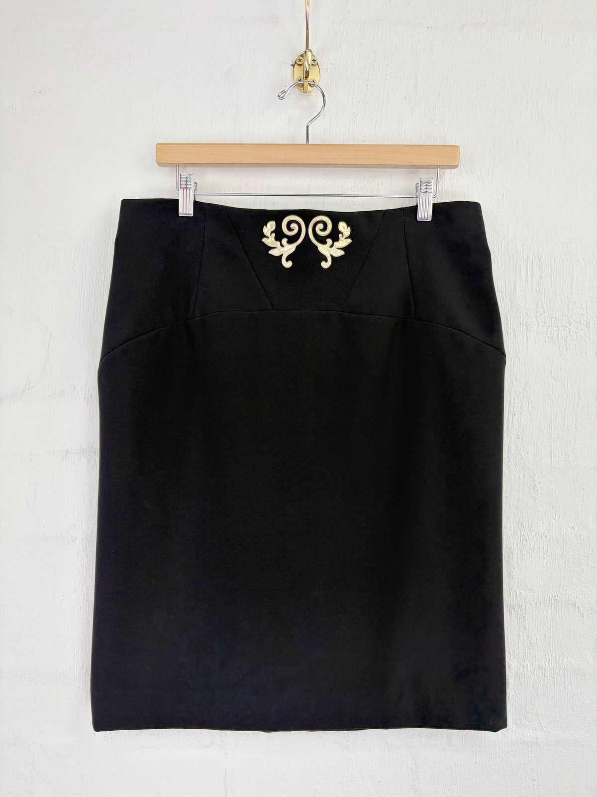 A black pencil skirt with a gold embroidered scroll motif at the waistband, hanging from a wooden hanger against a white textured wall.