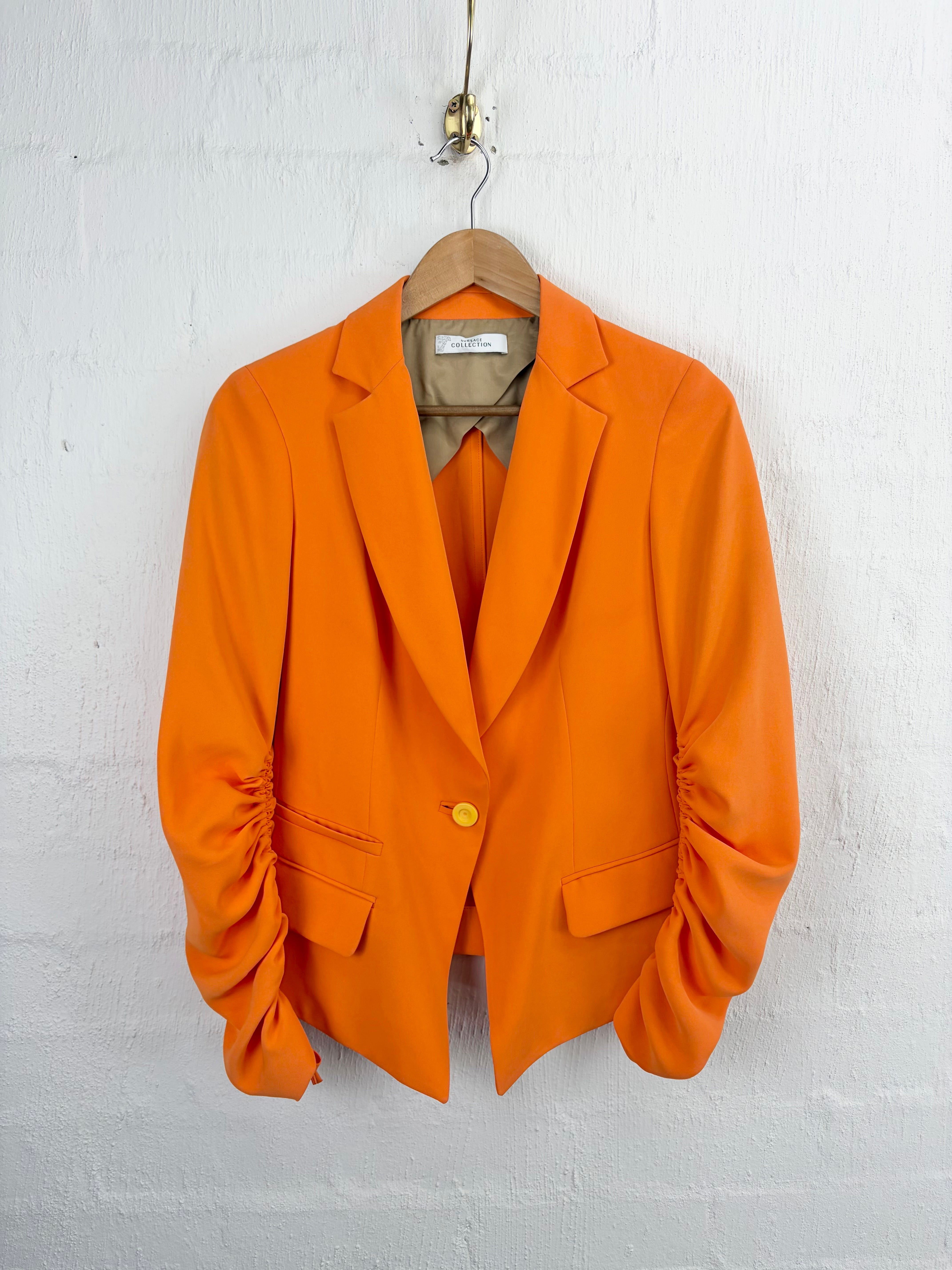 Bright orange tailored blazer with gathered sleeves and a single gold-tone button, hanging on a wooden hanger against a white painted brick wall.