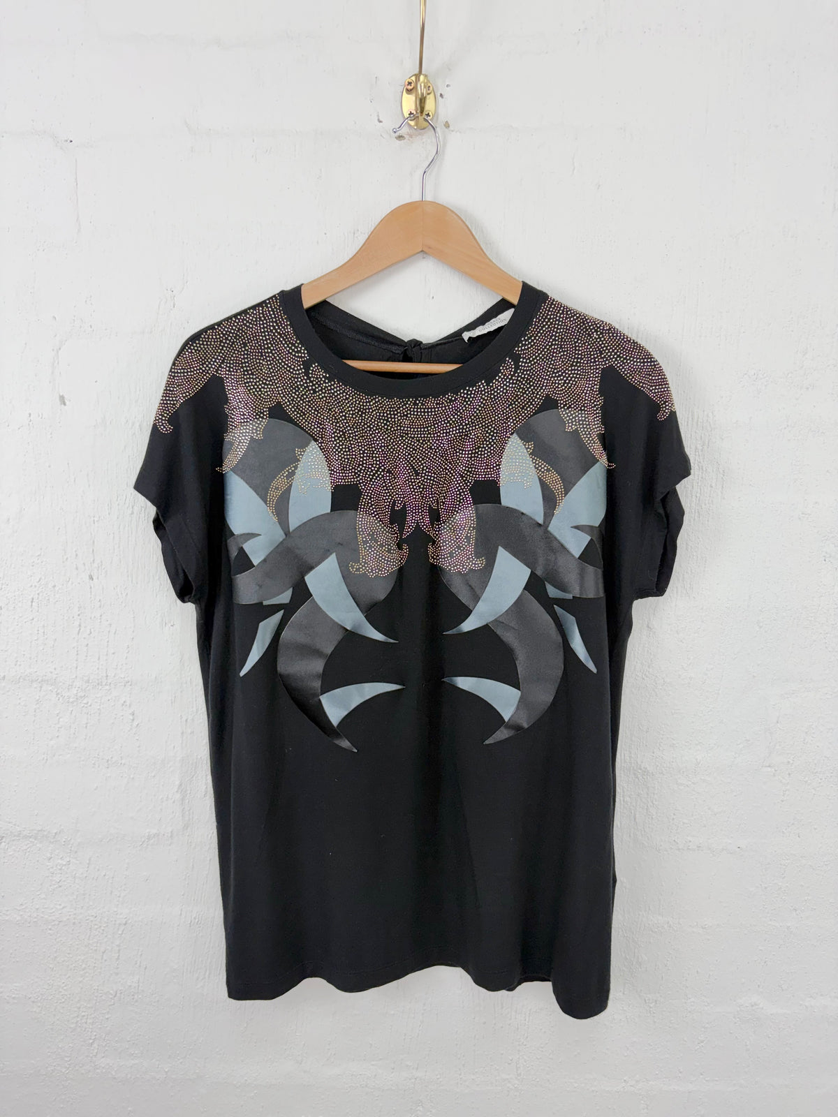 A black short-sleeve t-shirt with a large abstract butterfly-like graphic in gray, black, and metallic rose-gold on the front, hanging on a wooden hanger against a white painted brick wall.
