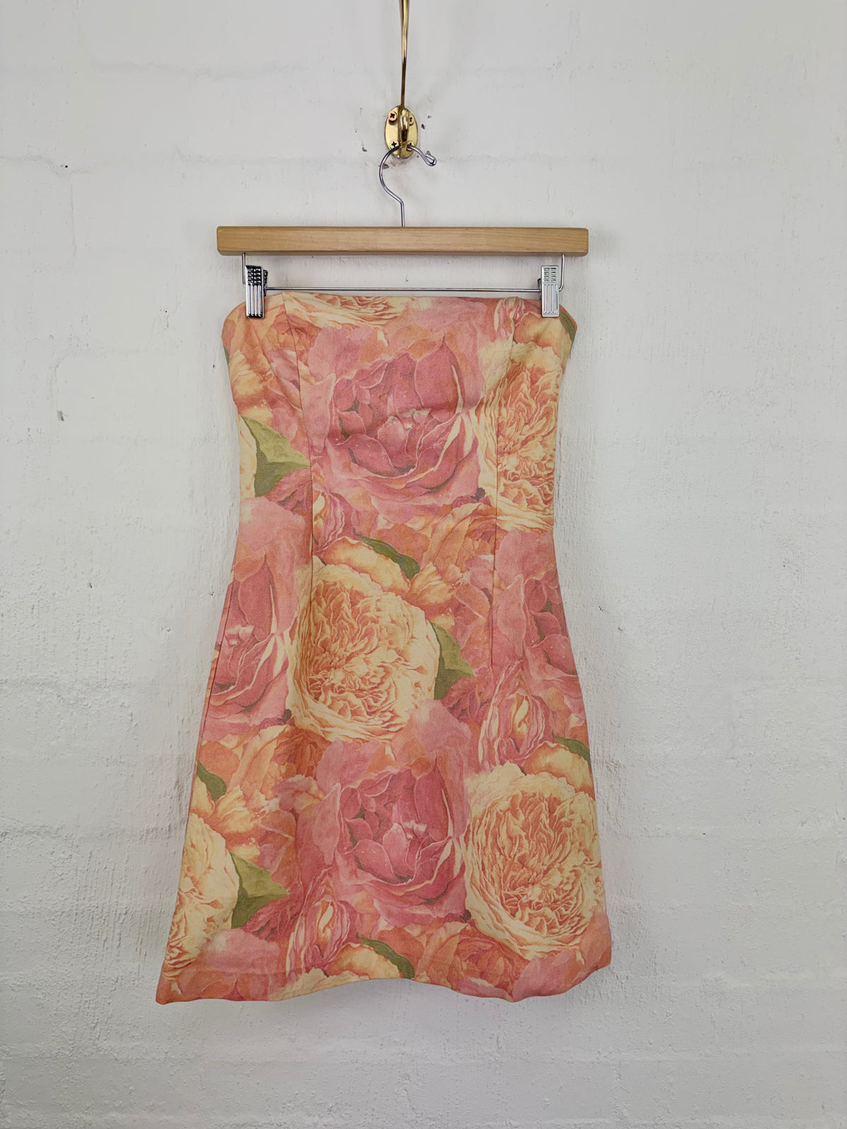 A strapless A-line dress with a vintage rose floral print in shades of pink, peach, and yellow, hanging from a wooden hanger against a white painted brick wall.