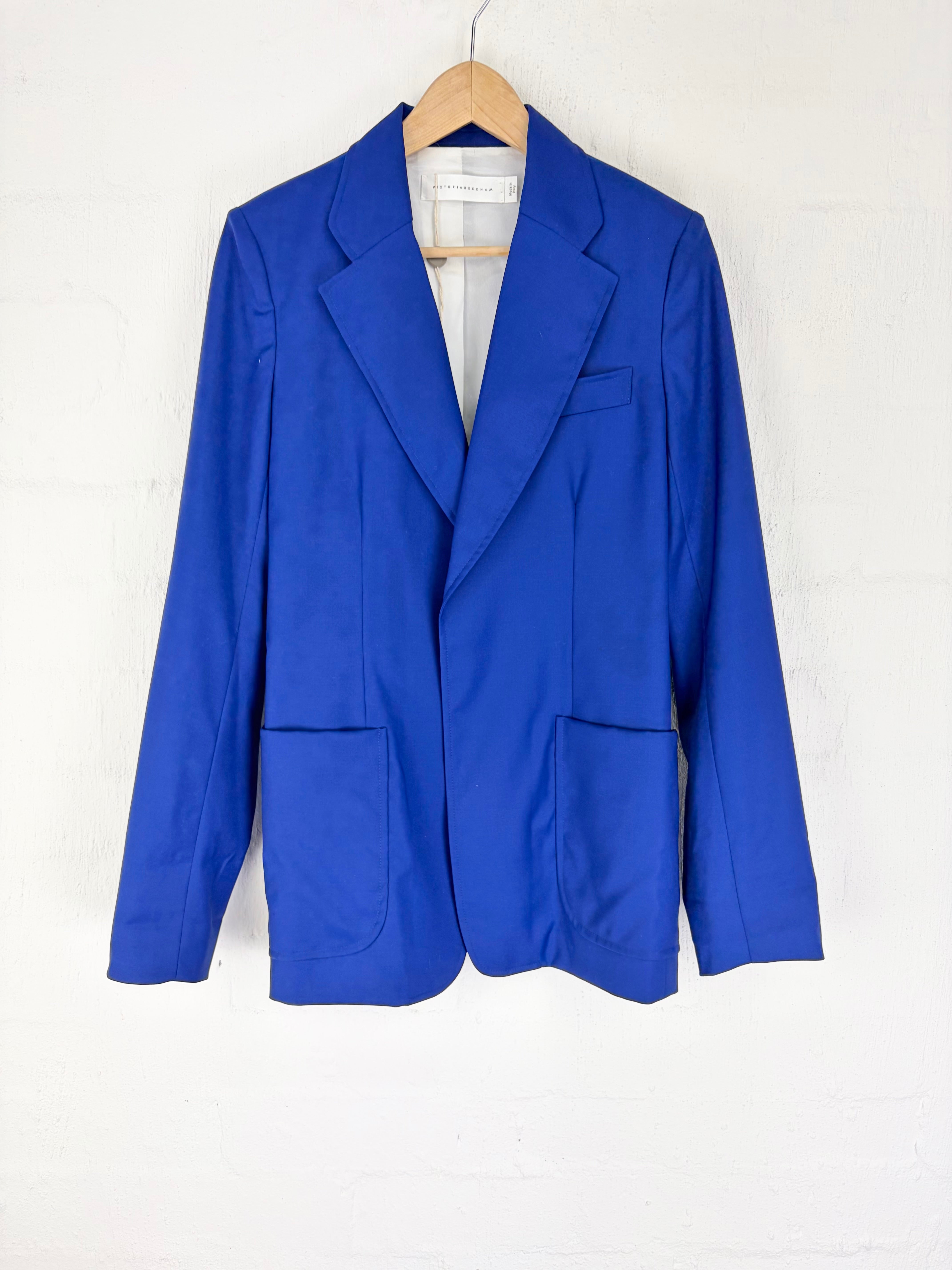 A bright royal blue blazer jacket hanging on a wooden hanger against a white painted brick wall.