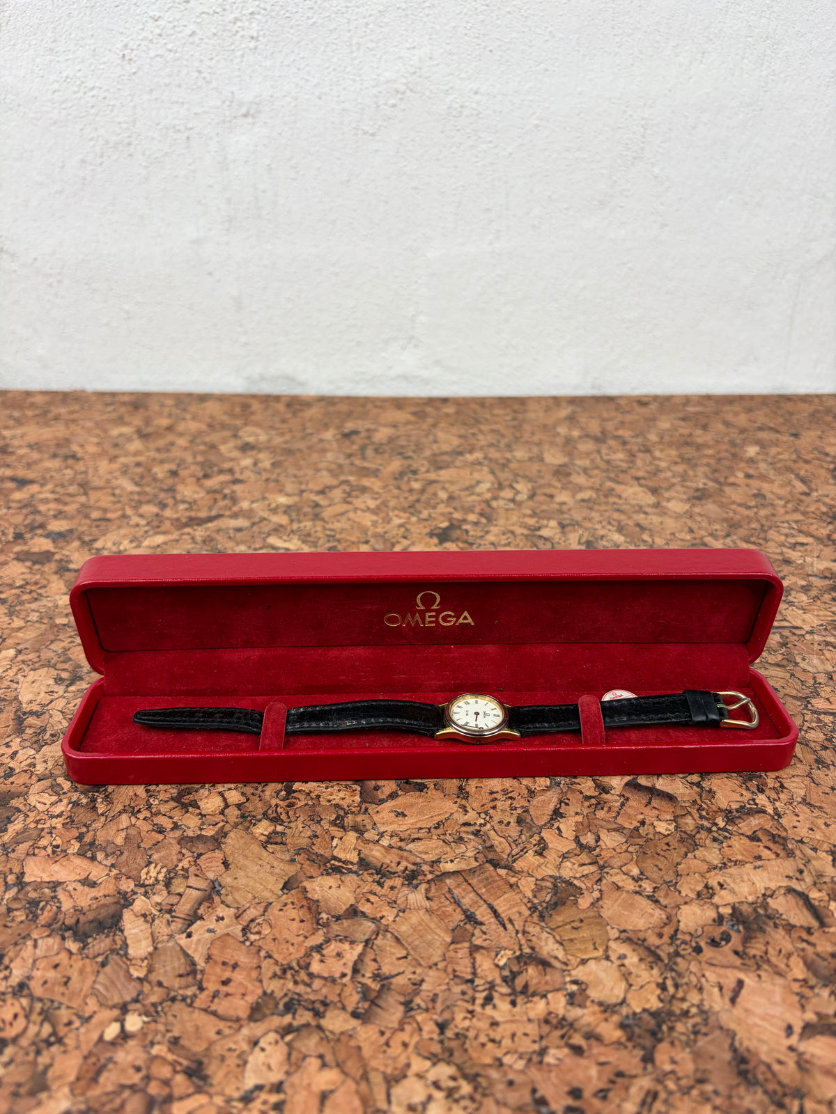 An Omega wristwatch with a black leather strap and gold-tone case, displayed inside its open red velvet Omega presentation box on a cork surface against a white wall.