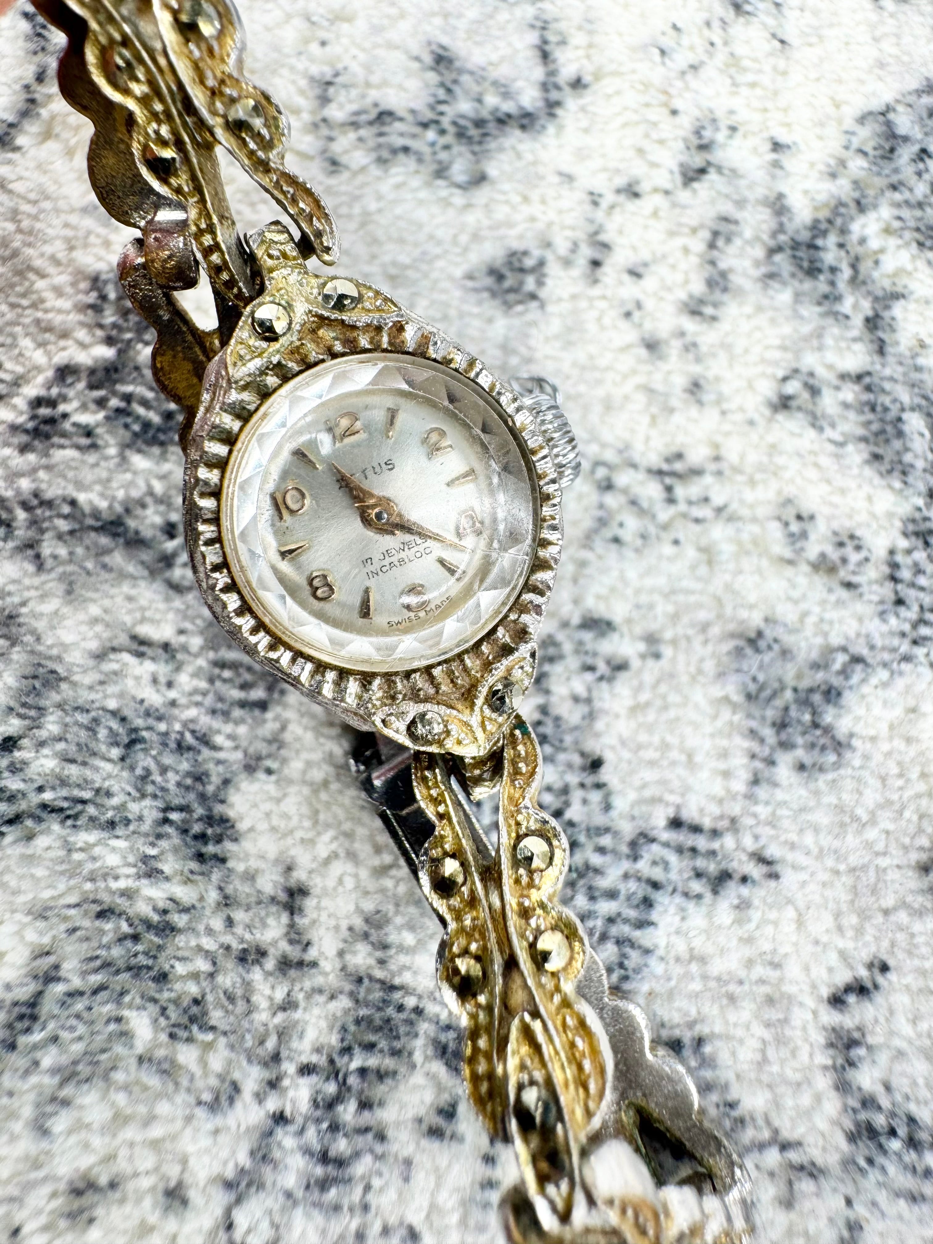 A vintage women's watch with an ornate gold and silver-tone link bracelet, resting on a textured gray and white fabric background.