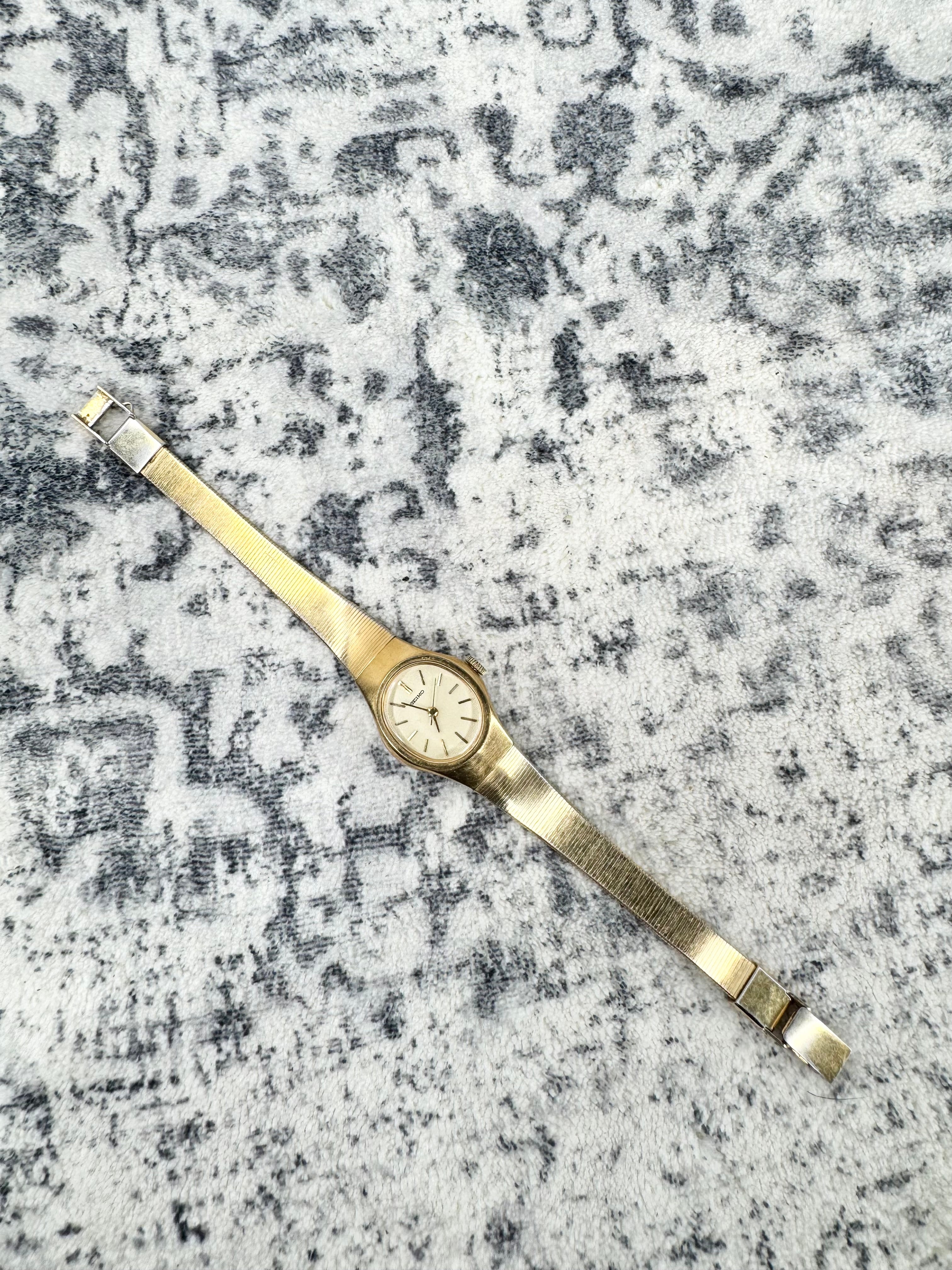 A vintage-style gold-tone wristwatch with a slim metal bracelet and a round dial, placed diagonally on a textured gray and white patterned rug.