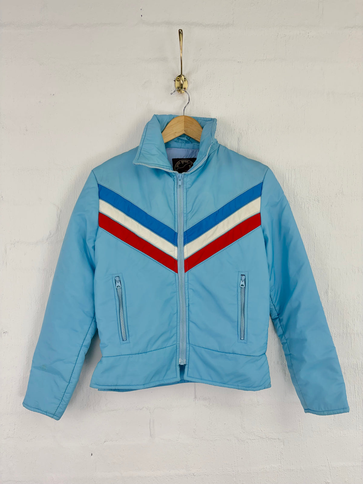 A light blue zip-up jacket with a red, white, and blue chevron pattern across the chest, hanging on a wooden hanger against a white brick wall.